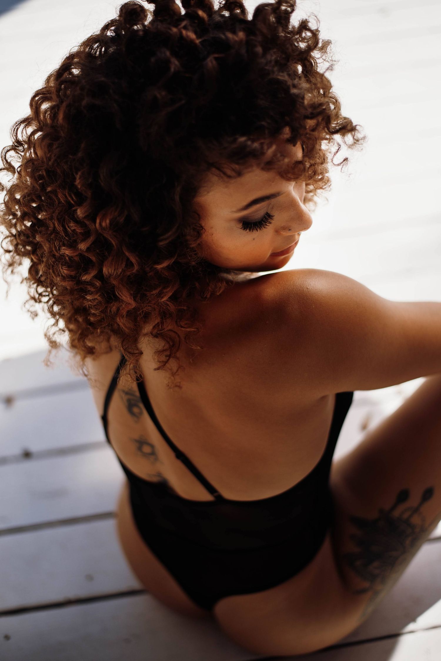 Woman with curly auburn hair and tattoos wearing black lingerie sitting on white wooden floor.