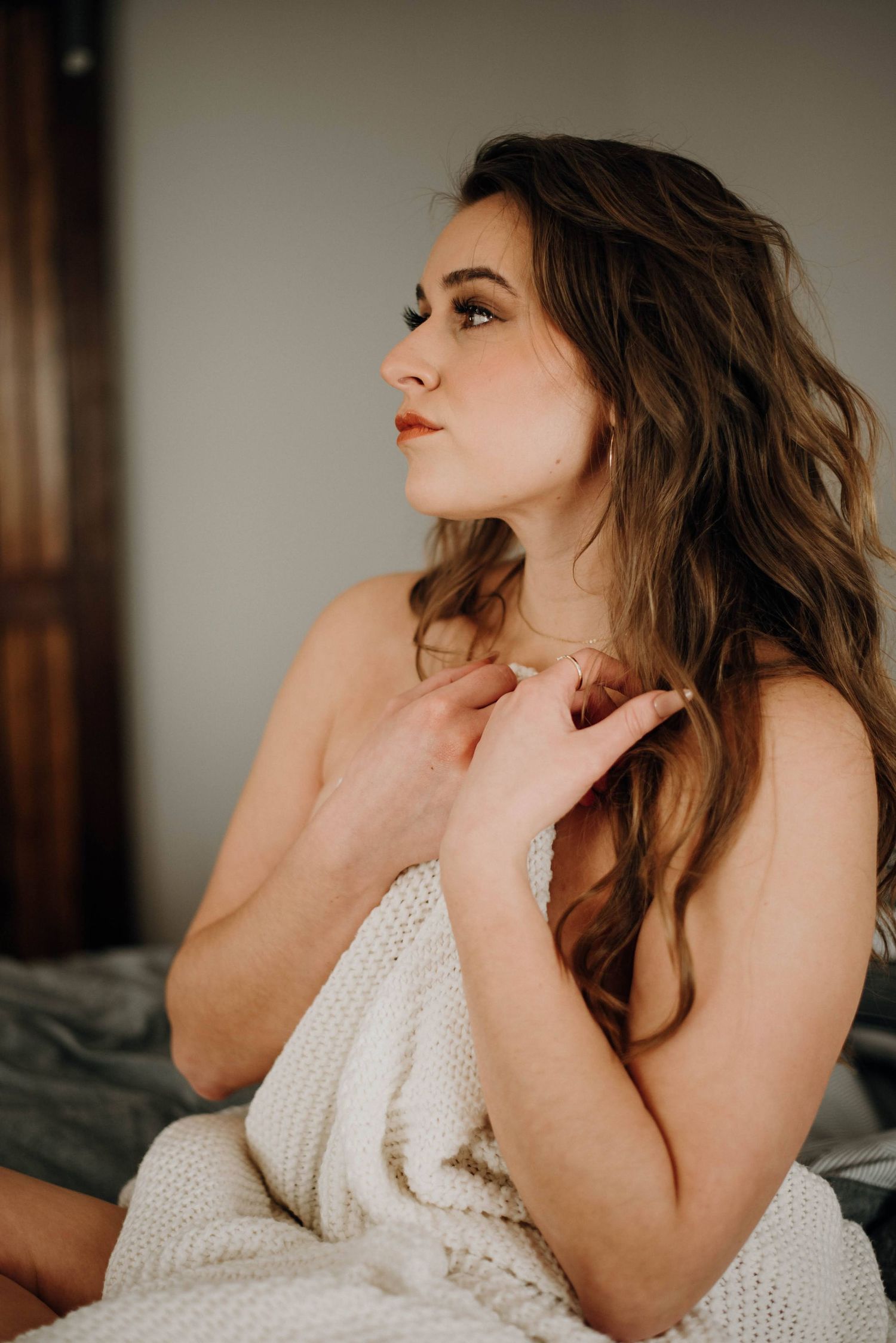 A young woman with long wavy brown hair wrapped in a white knit blanket sits on a bed, gazing upward thoughtfully.