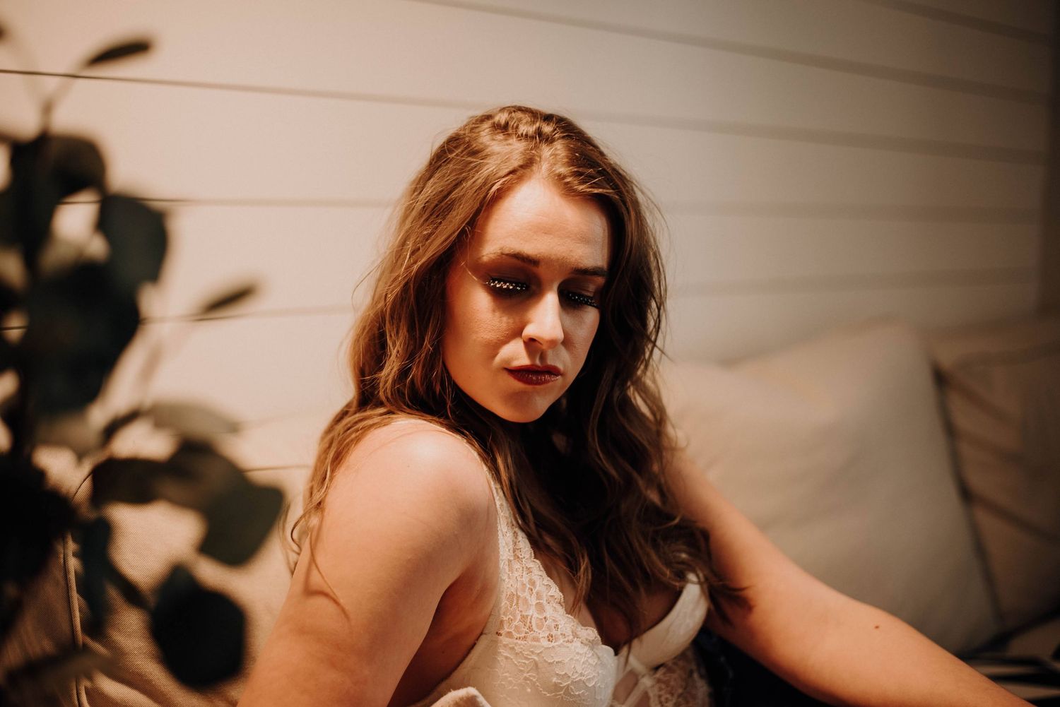 A young woman with long auburn hair in a lace top gazes downward in warm, moody lighting near a plant.