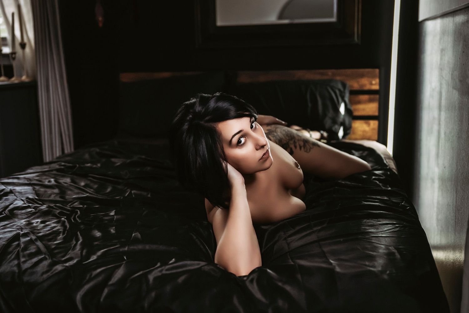 A woman with dark hair lies on black satin sheets in a moody, dimly lit bedroom setting.