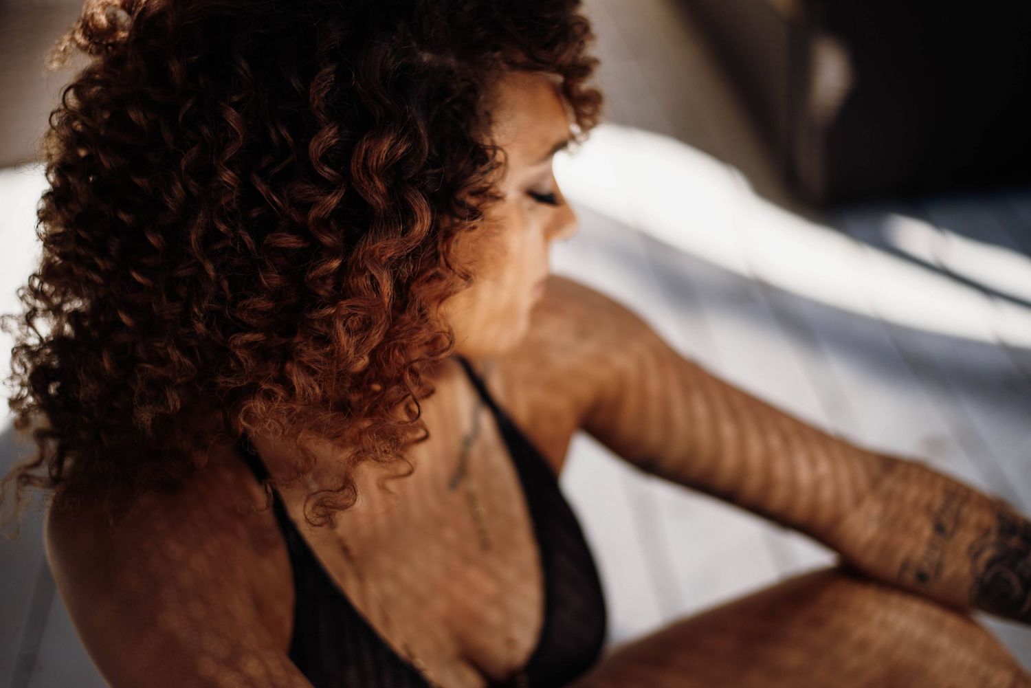 Woman with curly auburn hair in black lingerie sitting in sunlight.