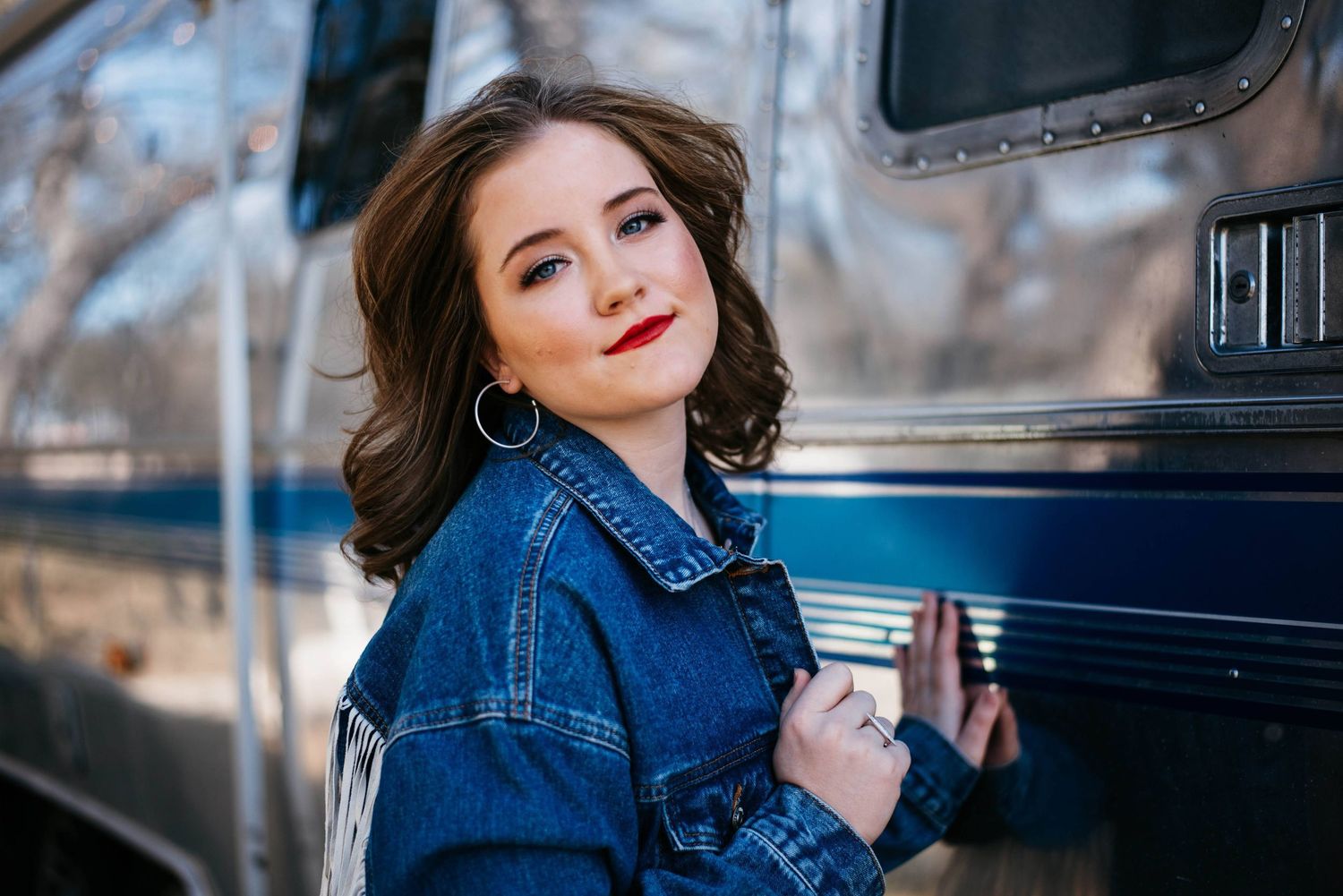 Young woman in a denim jacket with red lips posing confidently beside a vintage silver trailer outdoors.