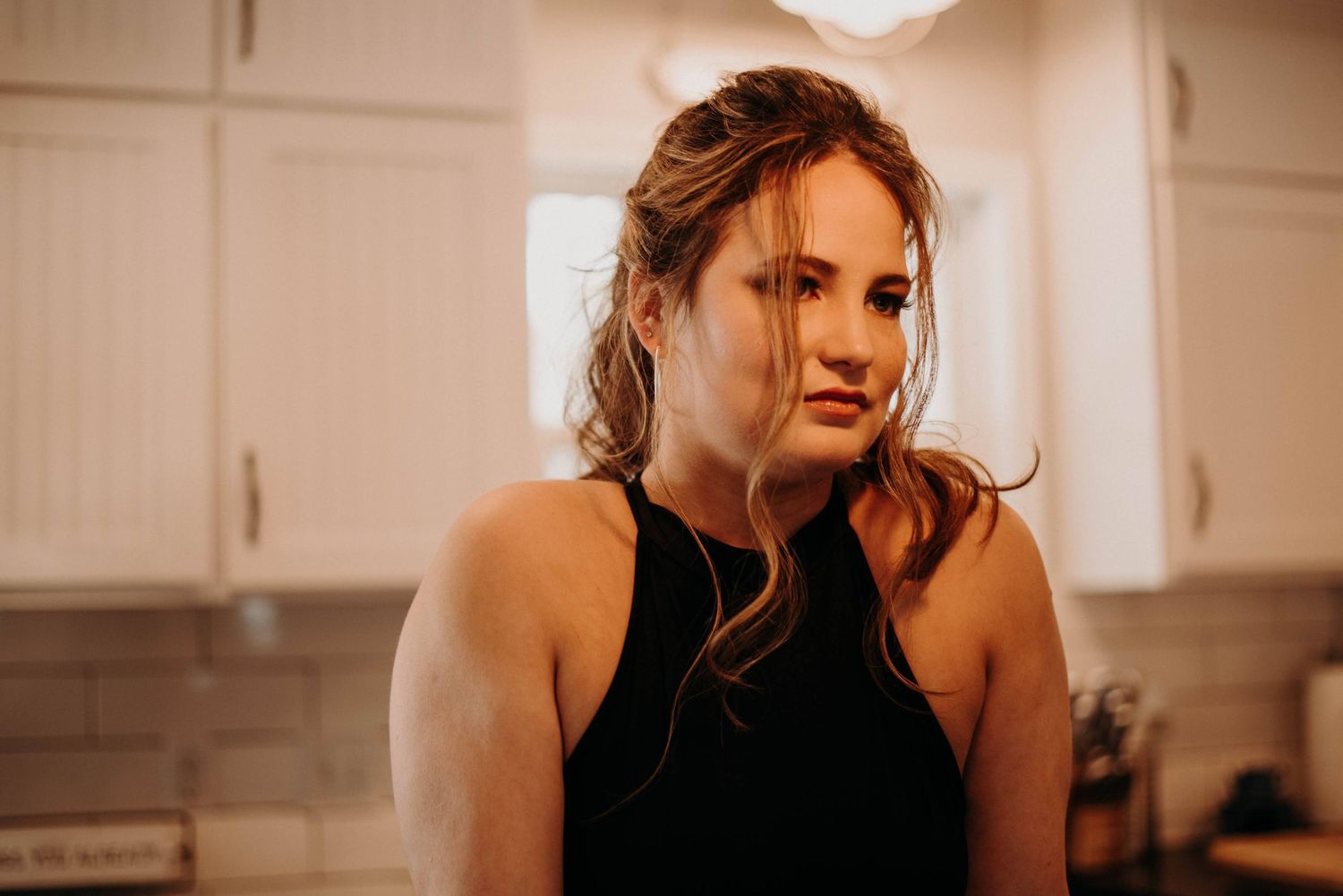 Young woman with wavy hair wearing a black tank top standing in a bright modern kitchen.