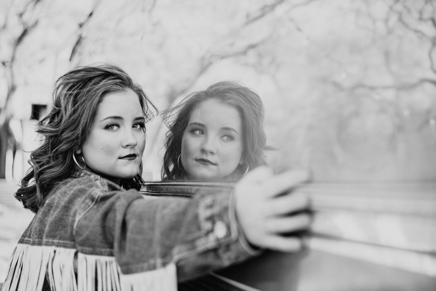 Black and white photo of a young woman taking a selfie with her reflection visible in a car window outdoors.