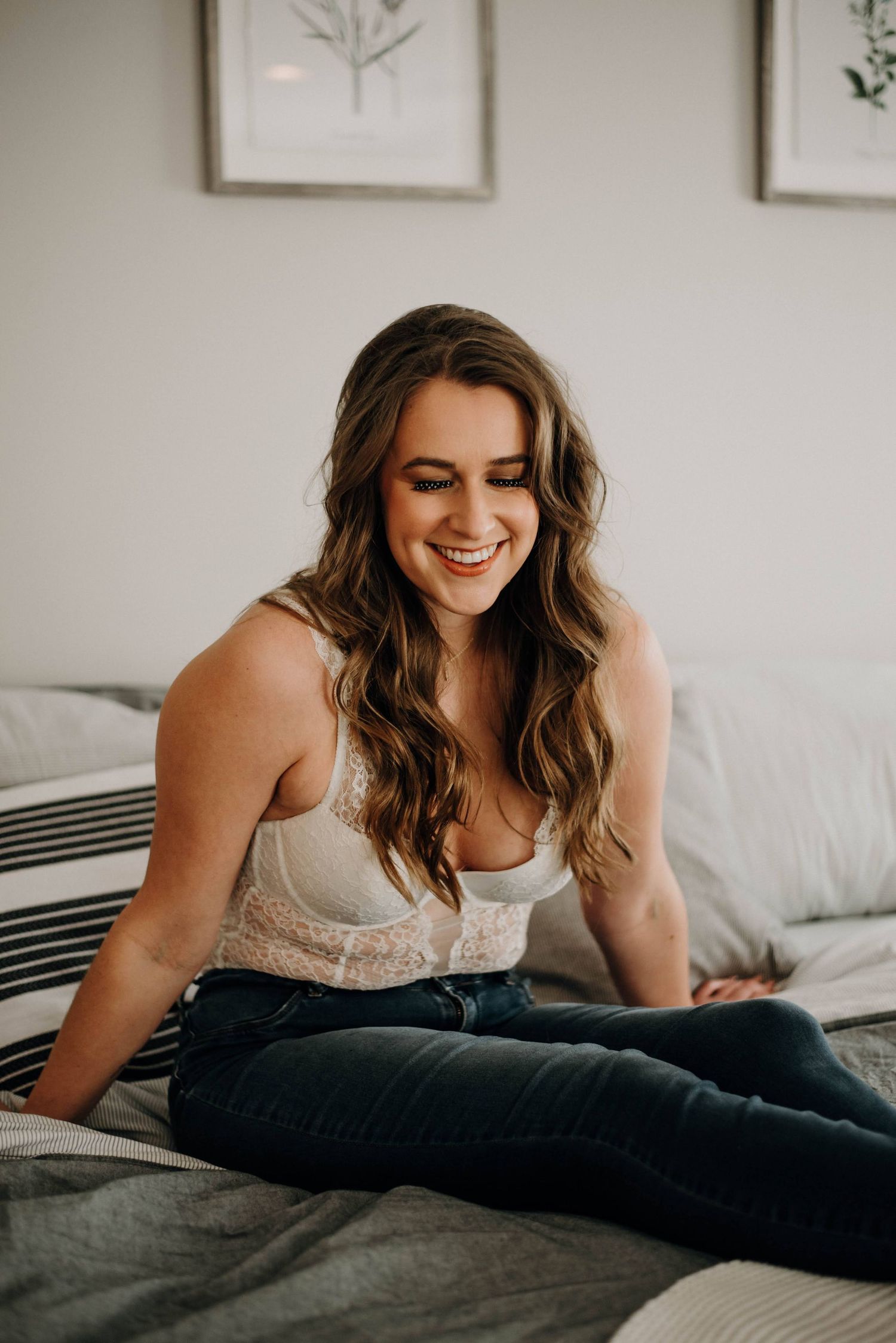 A smiling young woman with wavy brown hair sits cross-legged on a bed wearing jeans and a white lace top.