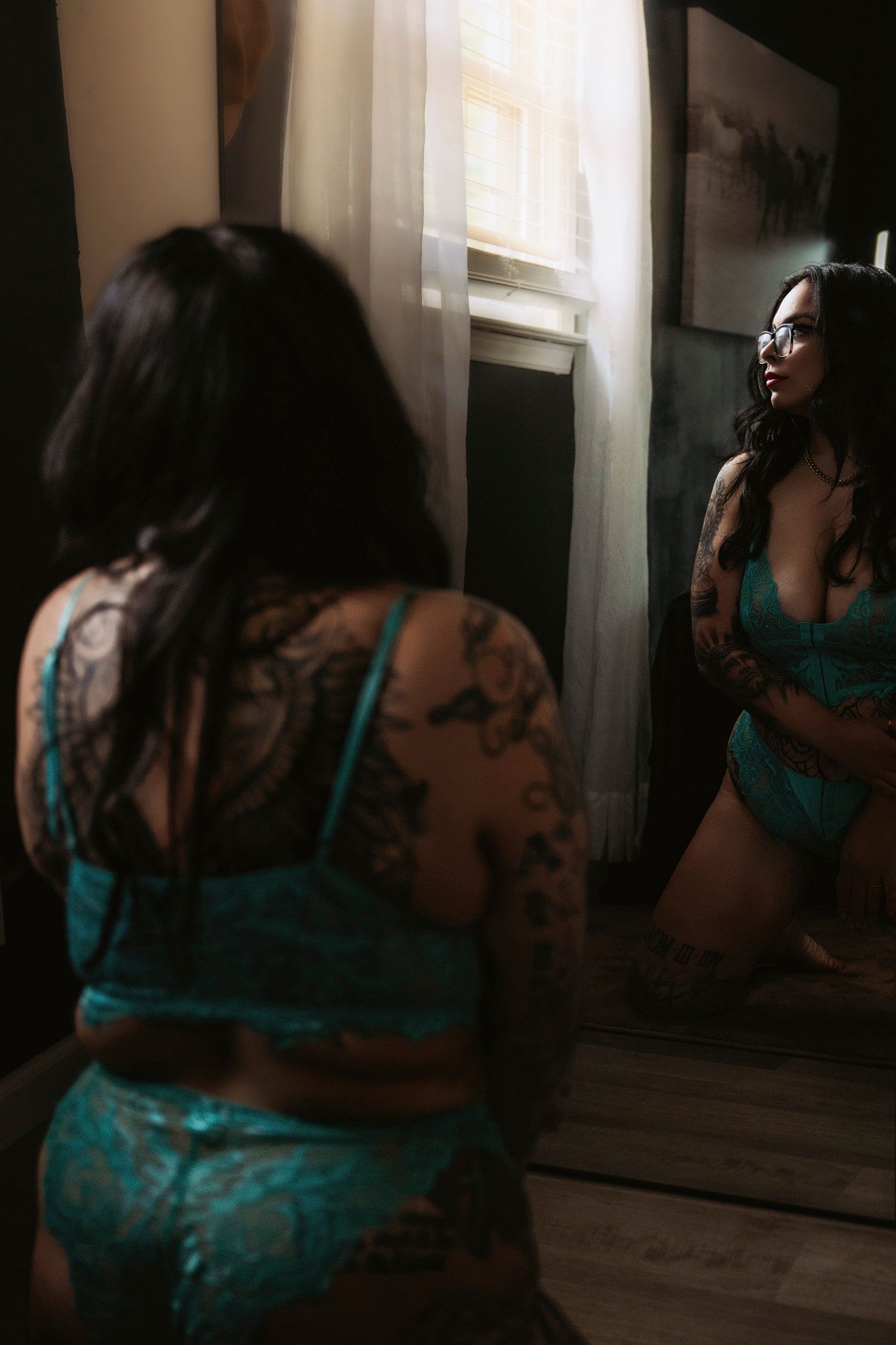 Tattooed woman in teal lace lingerie kneeling before a mirror in moody natural window light.