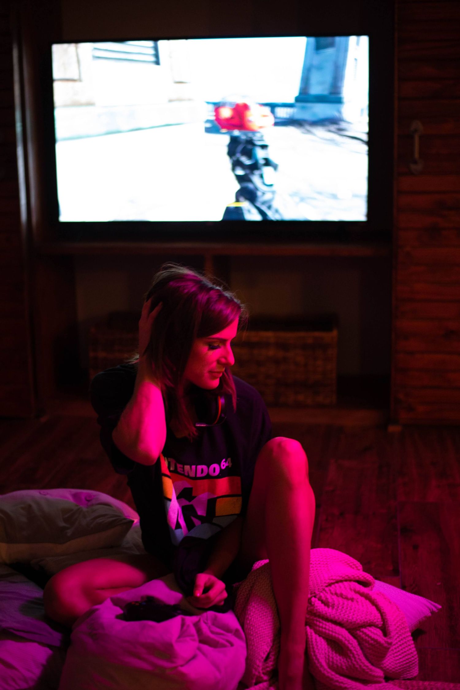 Young woman sitting on bed playing video games with pink lighting and a first-person shooter game on a large screen behind her.