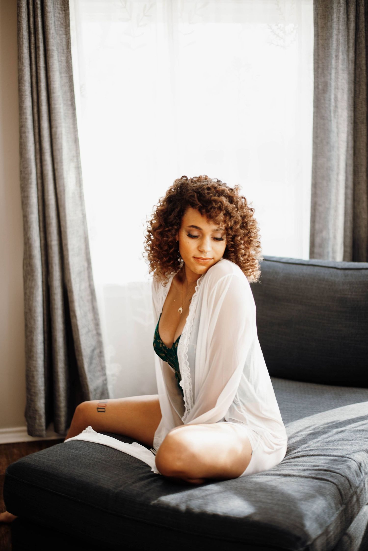 Curly-haired woman in white robe and green lingerie sitting on dark sofa by window.