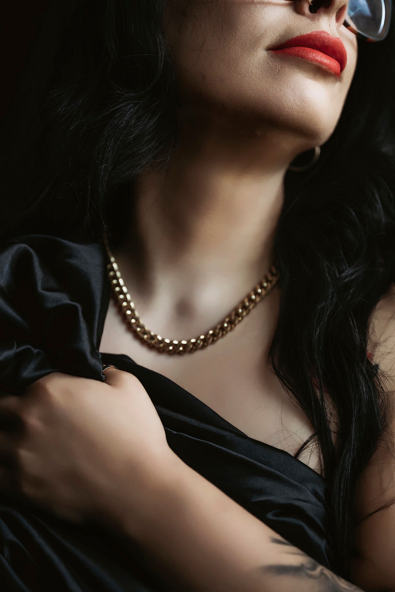 Woman in black satin dress with gold chain necklace, red lips, tattoo, and silver glasses in dramatic lighting.