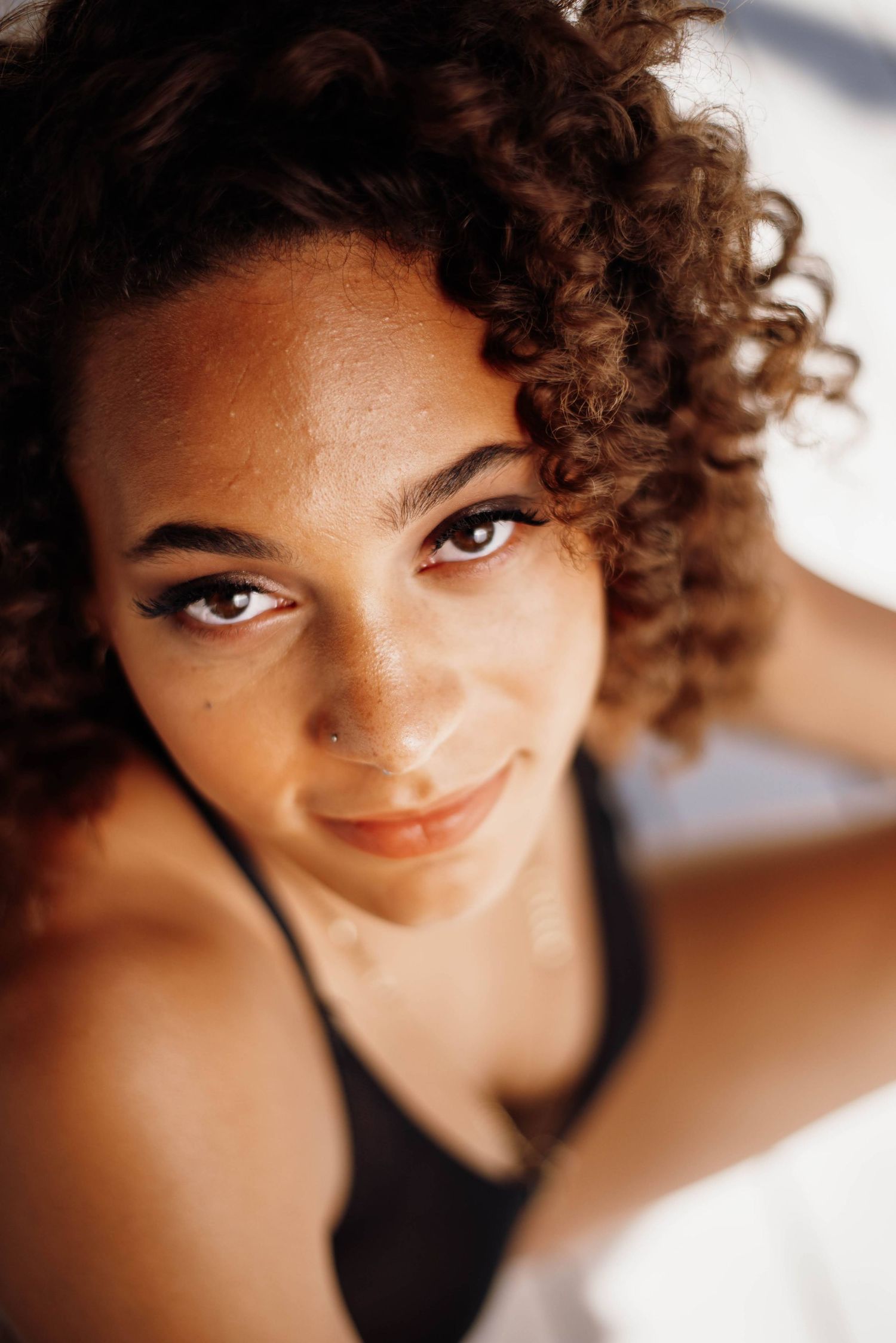 Young woman with curly brown hair and brown eyes smiling softly at camera in natural light.