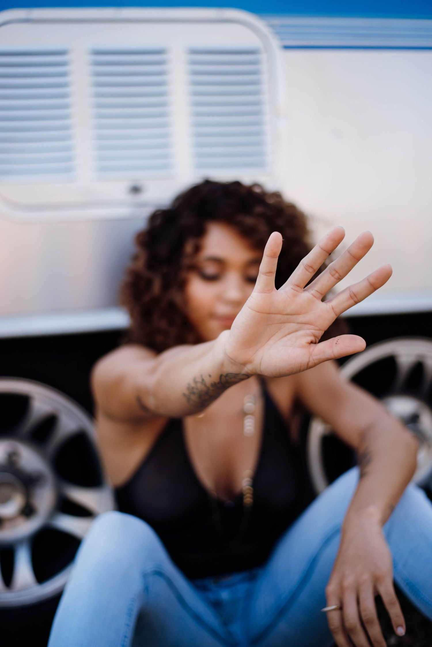 A young woman with curly hair sits by a van, extending her tattooed hand toward the camera.