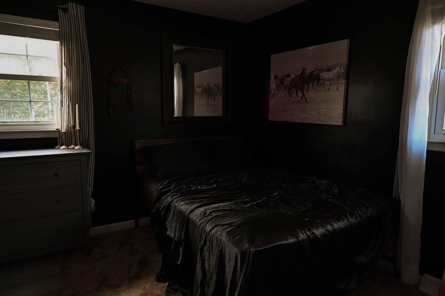 Dark bedroom with black walls, satin bedding, mirror, and horse artwork above bed near curtained windows.