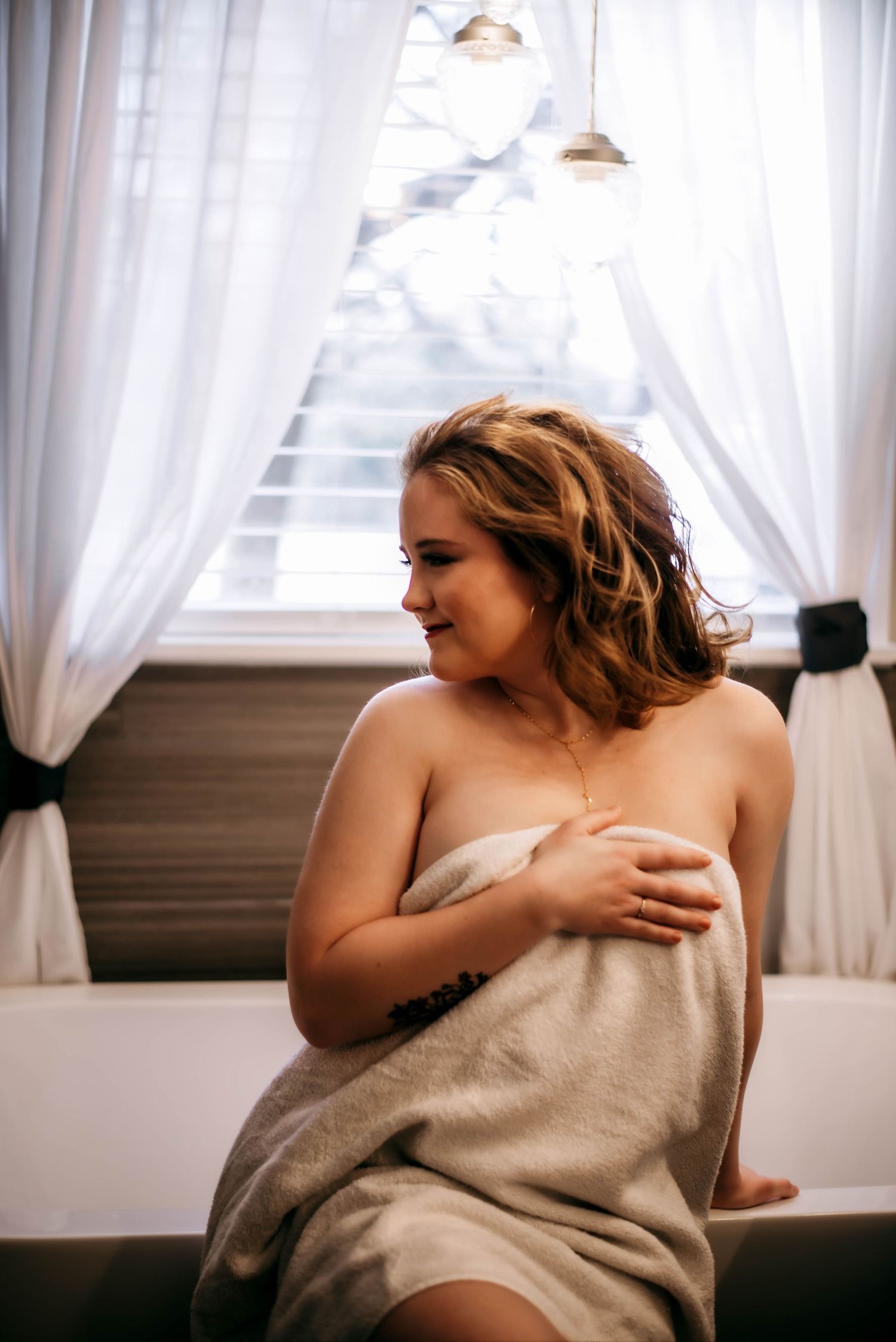 Woman wrapped in a towel sitting on the edge of a bathtub near white curtained windows in a bathroom.