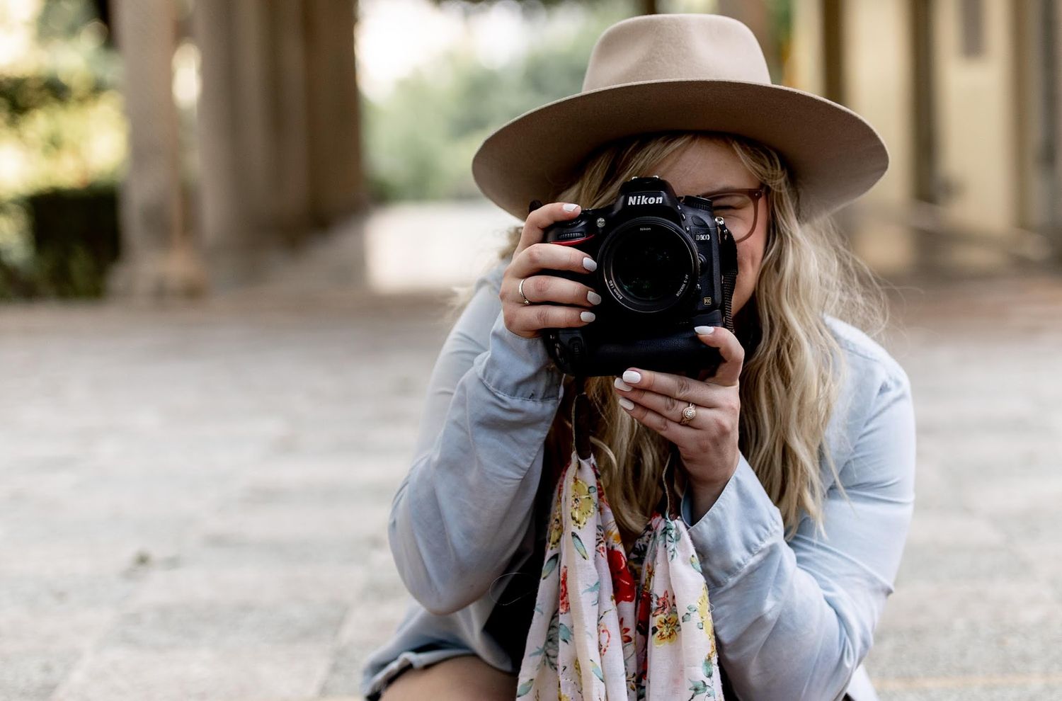 Blonde woman in wide-brim hat taking a photo with a Nikon camera while crouching outdoors.