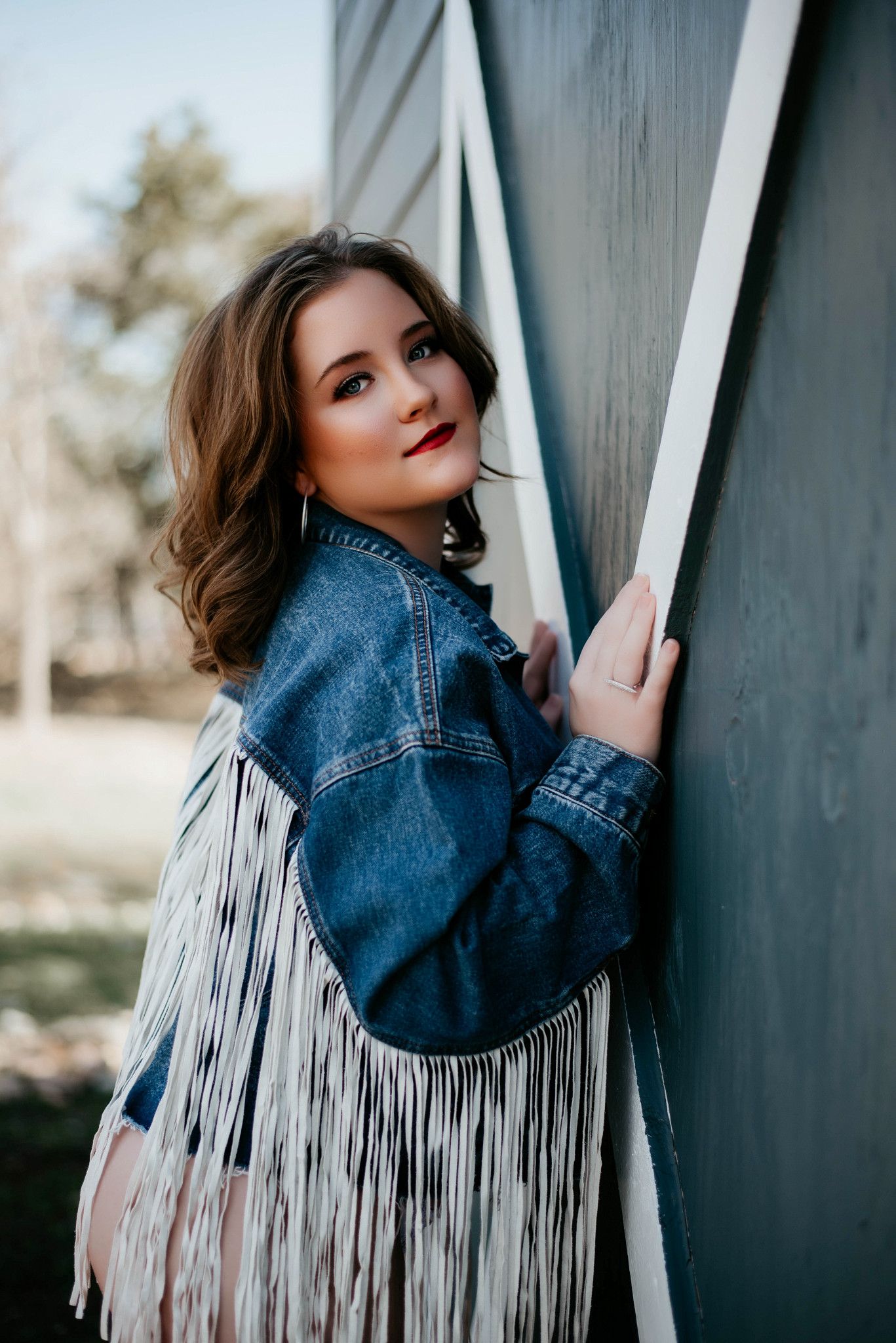 Young woman in fringed denim jacket posing against a teal barn wall outdoors.