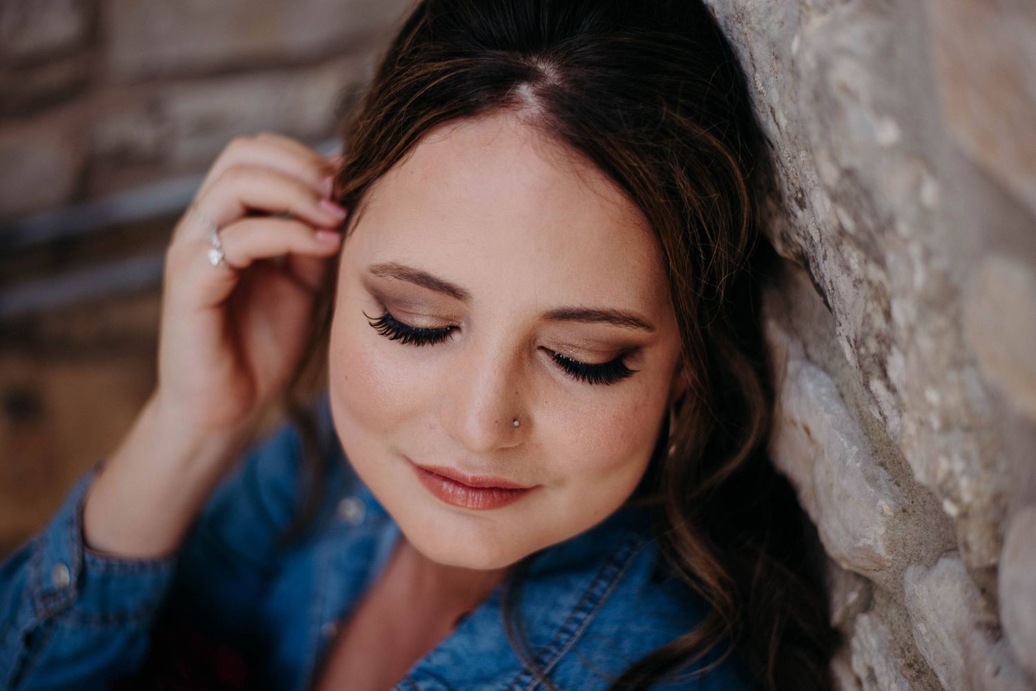 Close-up portrait of a brunette woman with elegant makeup leaning against a stone wall in a denim jacket.