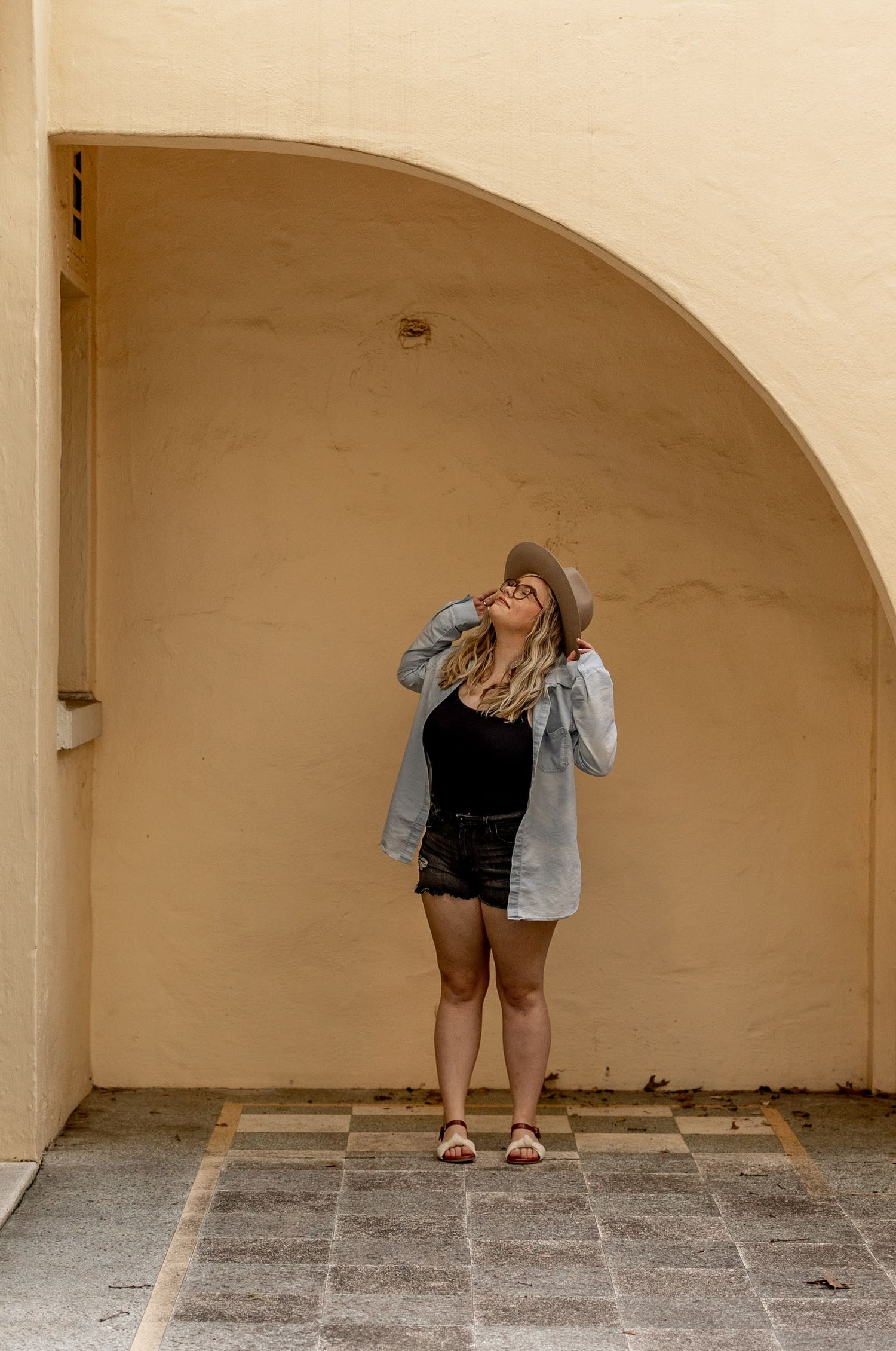 Woman in denim jacket, black shorts, and hat posing under an arched beige wall outdoors.