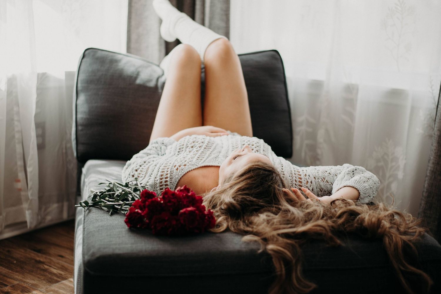 Woman lying on a gray sofa with red roses, wearing a white knit top, long blonde hair cascading down.