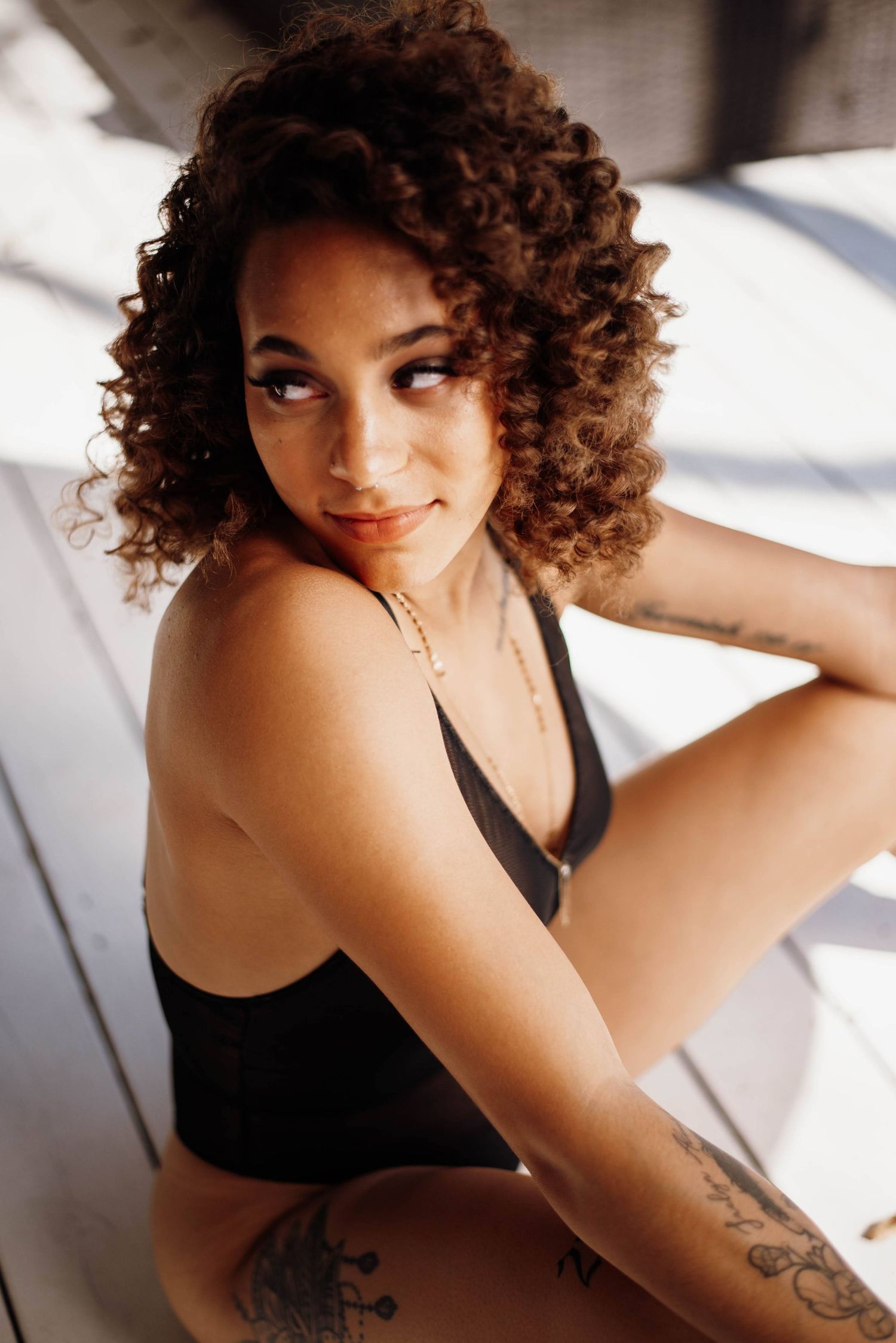 Young woman with curly hair and tattoos poses in a black swimsuit in natural light.