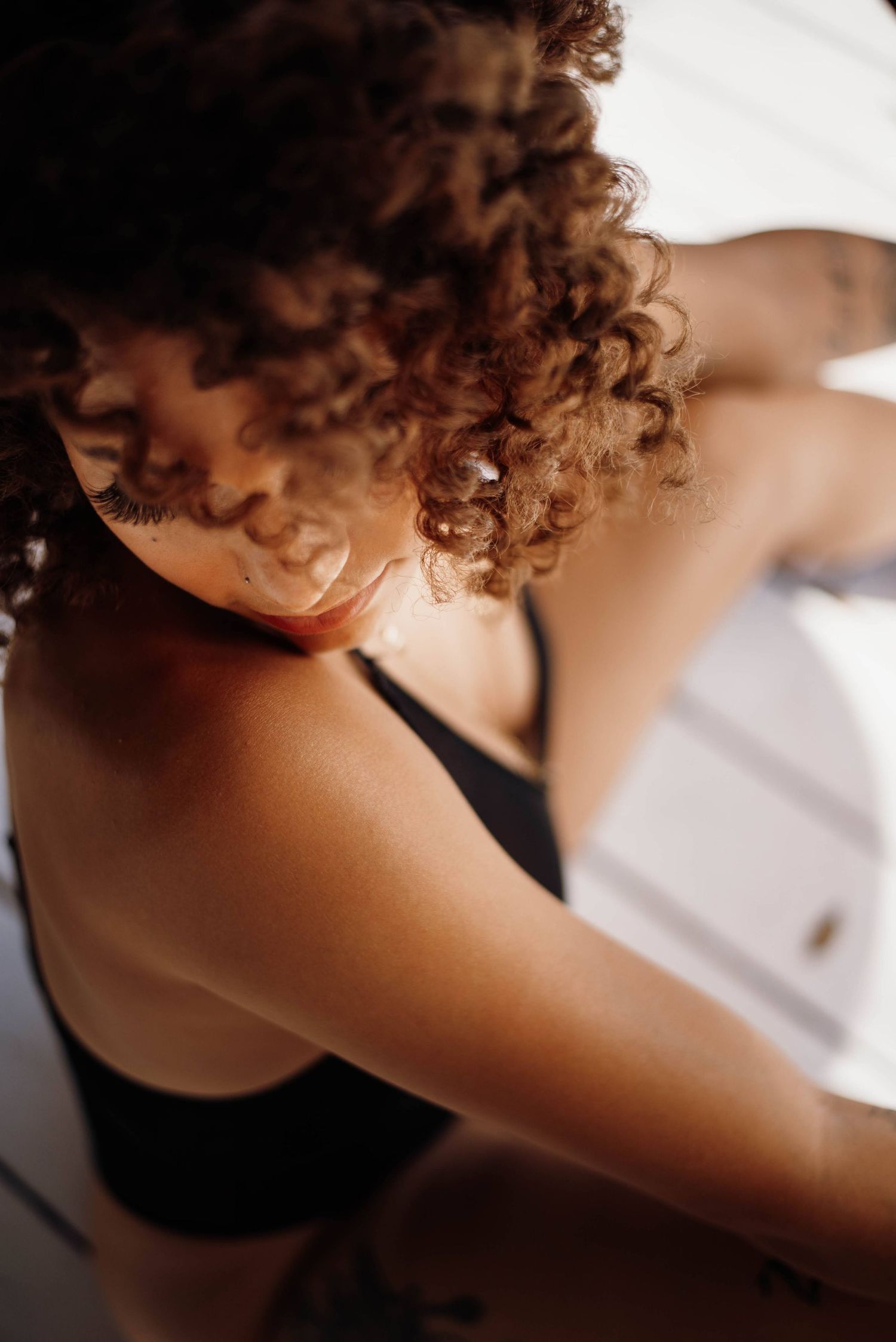 Close-up of curly-haired woman in black bralette with warm sunlight on her shoulder.
