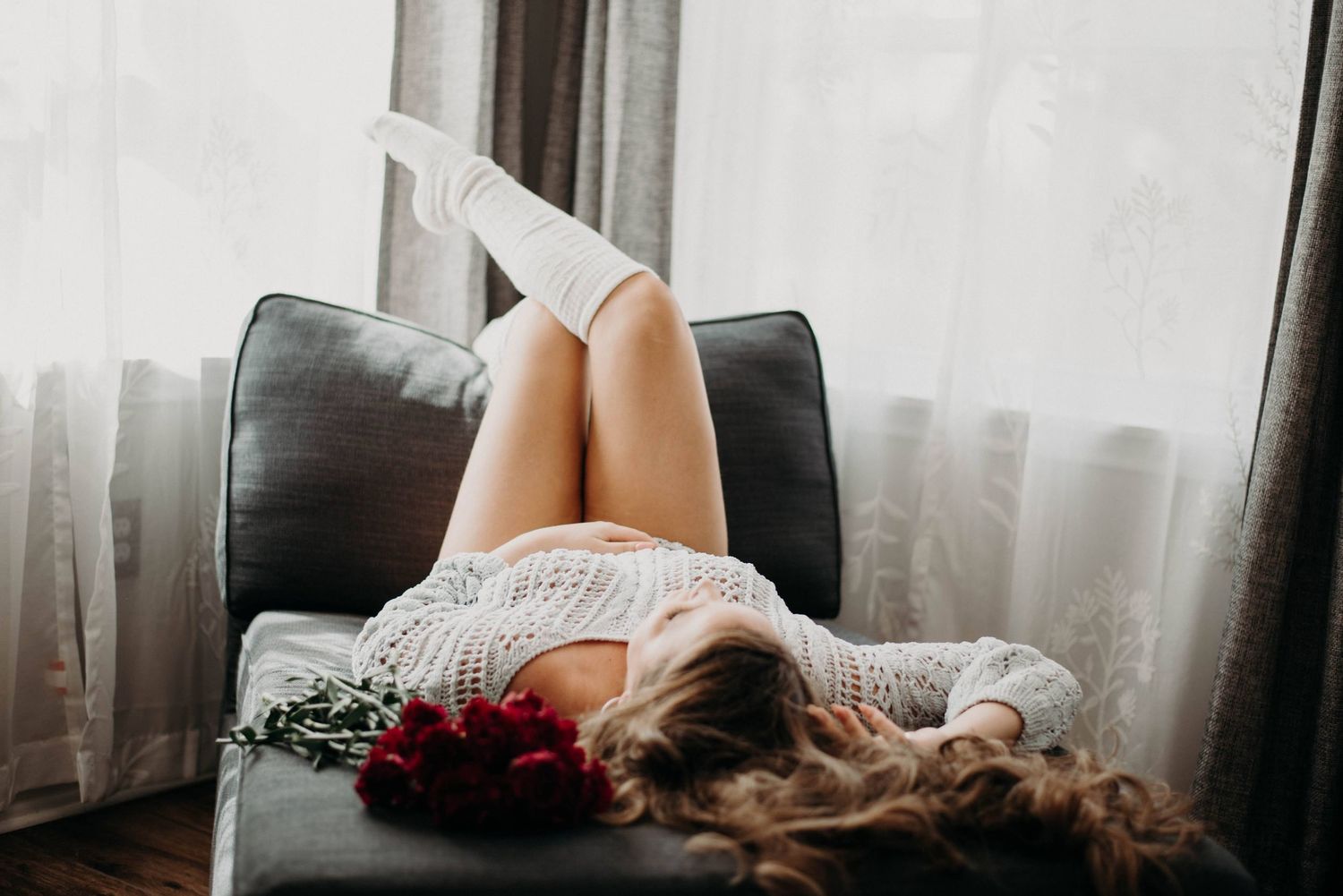 A woman in white knitwear and knee-high socks relaxes on a gray chair with red flowers near a bright window.
