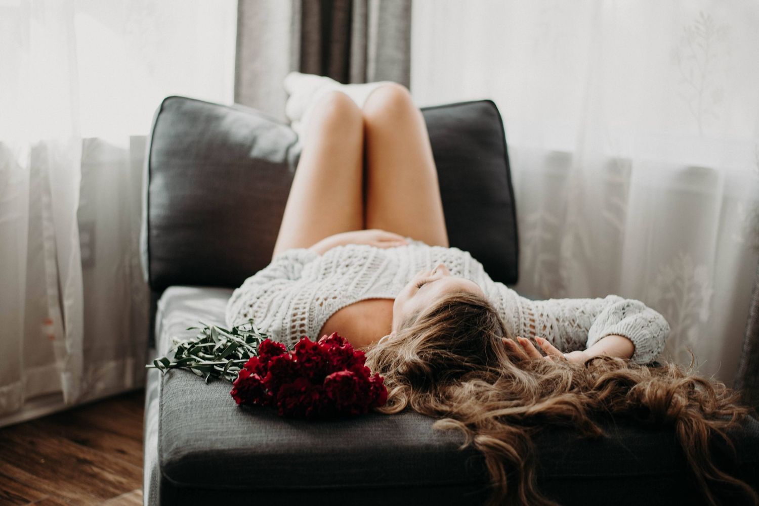 Woman in white knit dress lying on gray sofa with red roses, hair spread out, near sheer curtains.