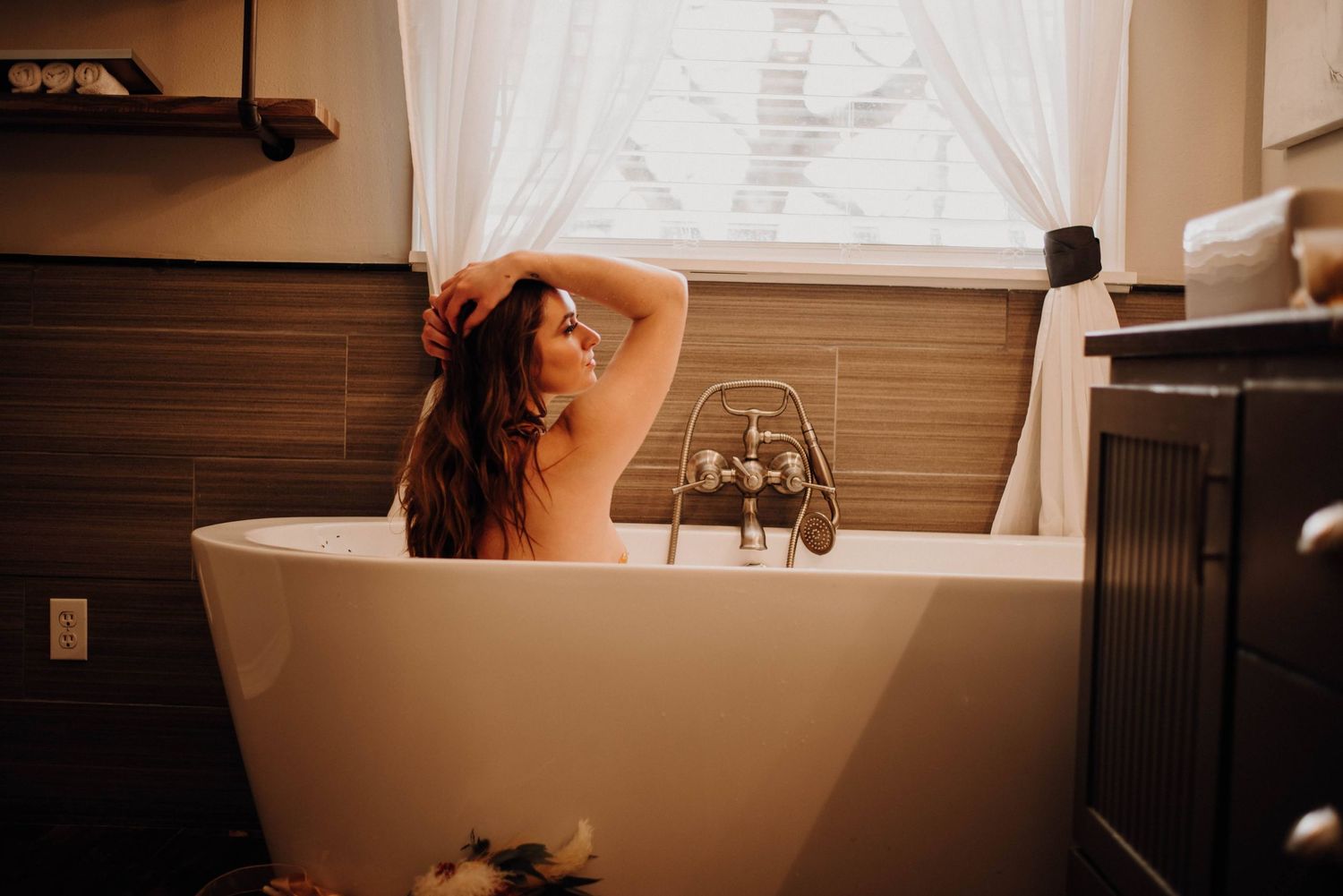Woman relaxing in freestanding soaking tub near window in elegant bathroom with warm lighting.