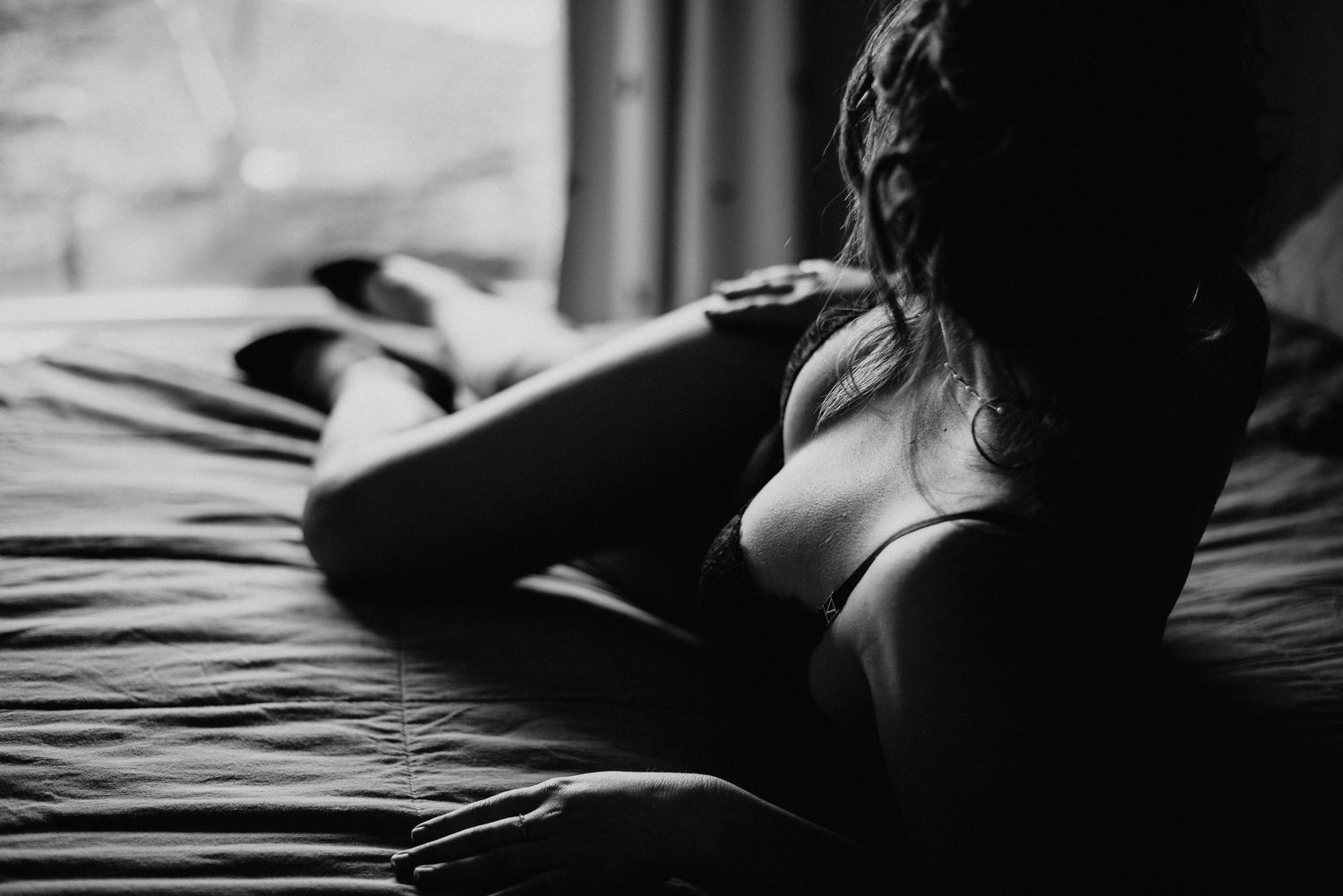 Black and white boudoir photo of a woman in lingerie posing on a bed near a window.