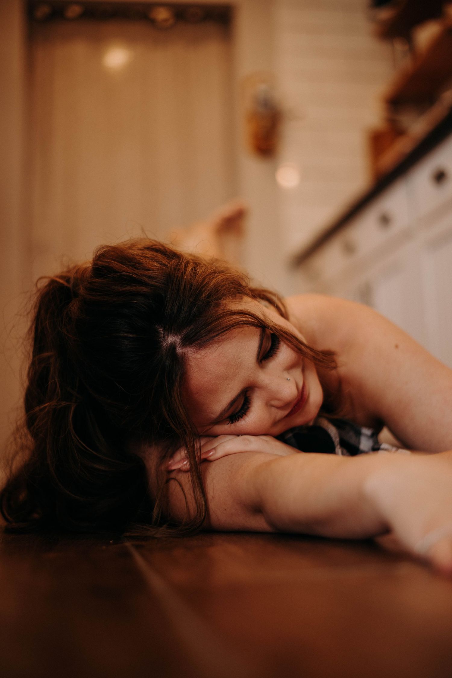 Woman with curly hair resting her head on a wooden table in a warmly lit kitchen.