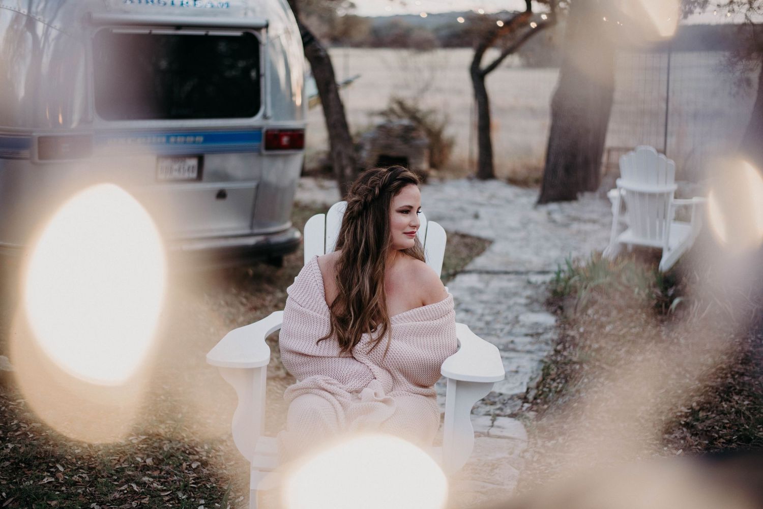 Bride in blush dress seated in white garden chair with bokeh lights and vintage van in background.