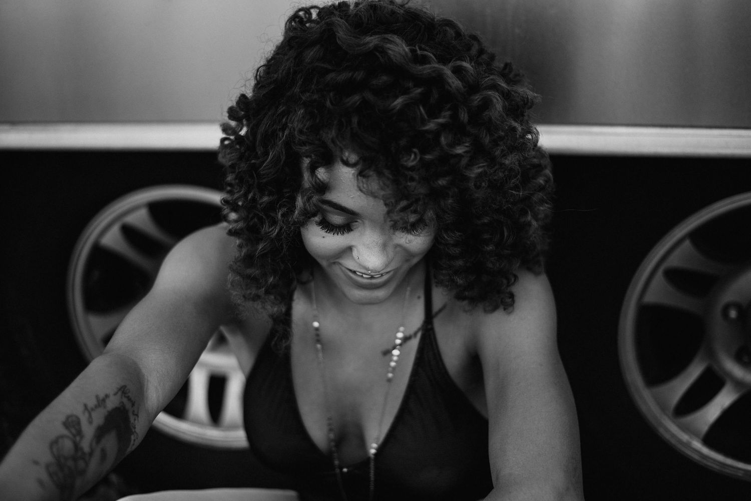 Black and white portrait of curly-haired woman with tattoo smiling downward in moody setting.