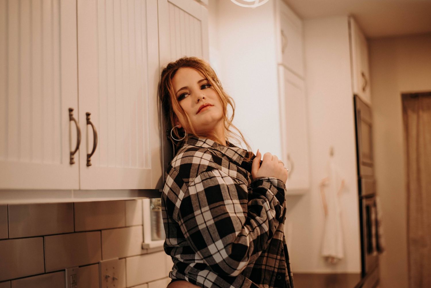Woman in plaid flannel shirt leaning against white kitchen cabinets with subway tile backsplash.