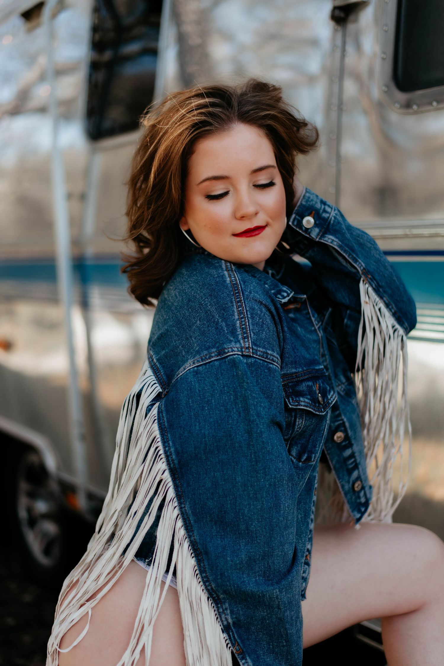 Young woman in fringed denim jacket poses outdoors near silver Airstream trailer with red lips and brown hair.