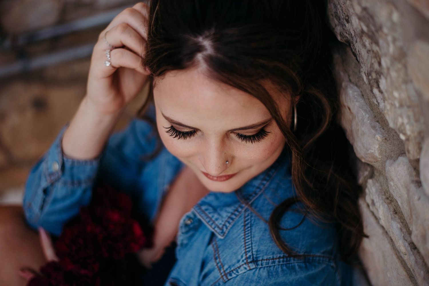 Close-up of brunette woman with dramatic lashes wearing denim jacket, leaning against stone wall.