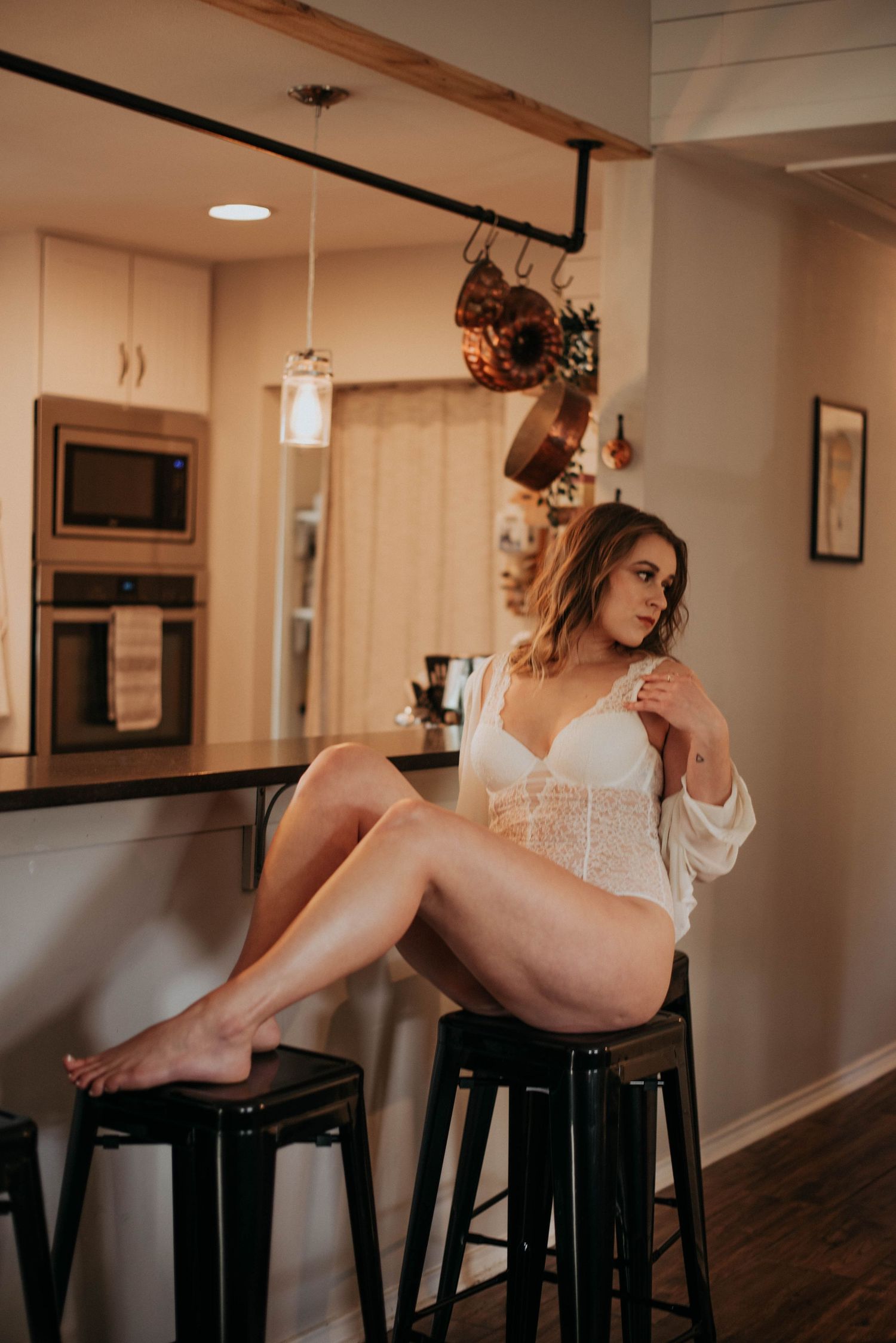 Woman in white lace bodysuit sitting on a barstool in a modern kitchen interior.