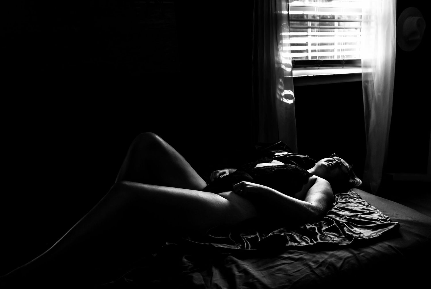 Black and white artistic photo of a person reclining on a bed near a window with light filtering through blinds.