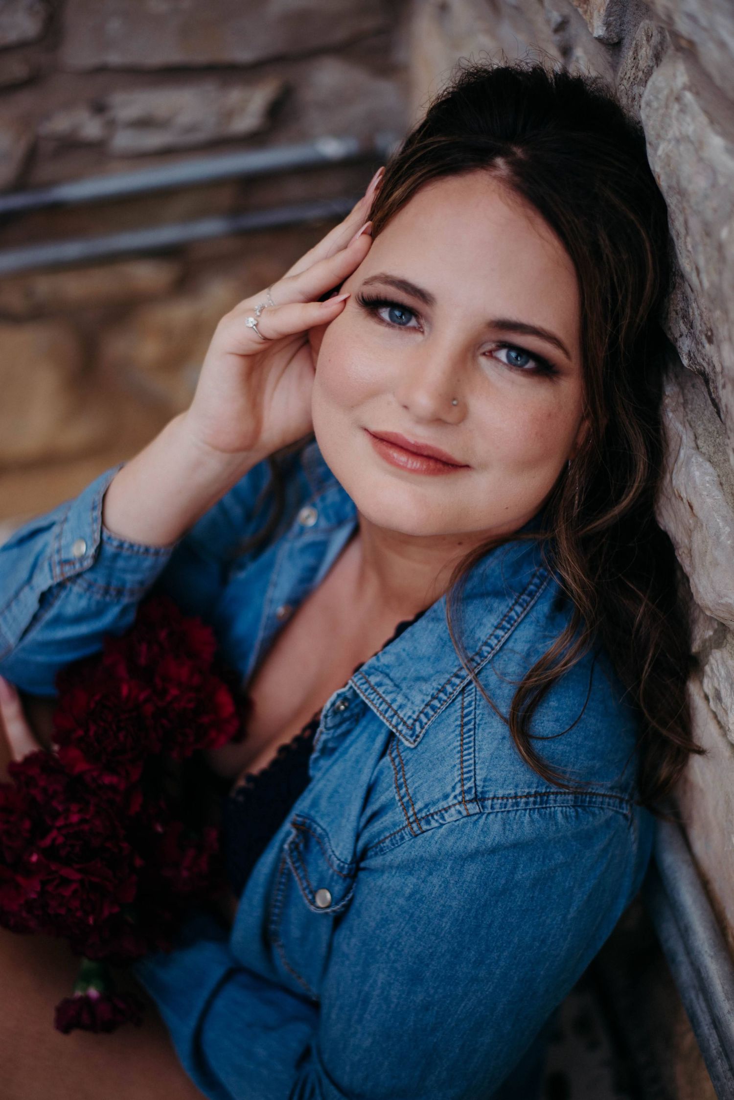 Young woman in denim shirt holding dark red flowers, leaning against stone wall, smiling with blue eyes.