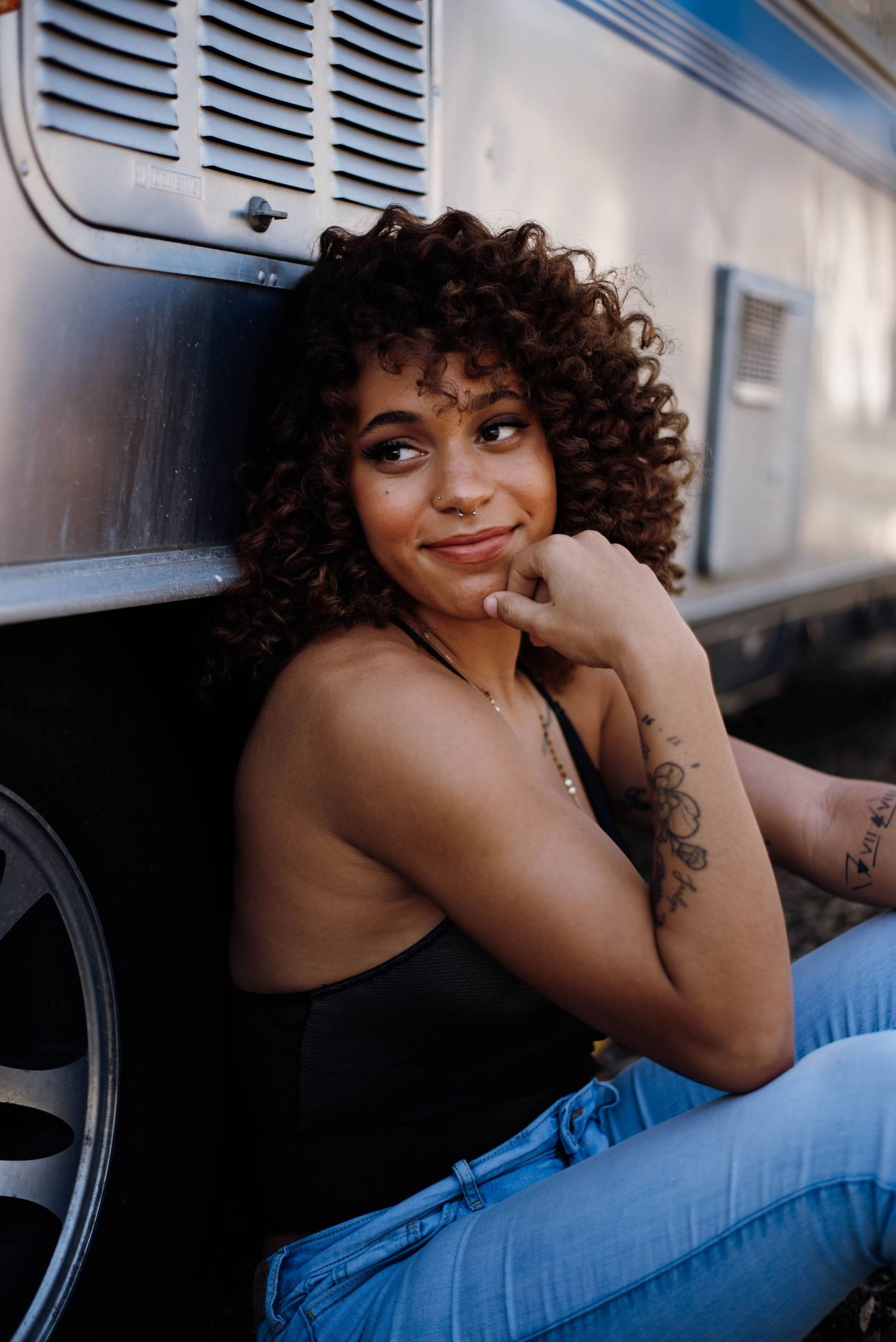 A curly-haired woman with arm tattoos sits against a silver van, smiling thoughtfully.