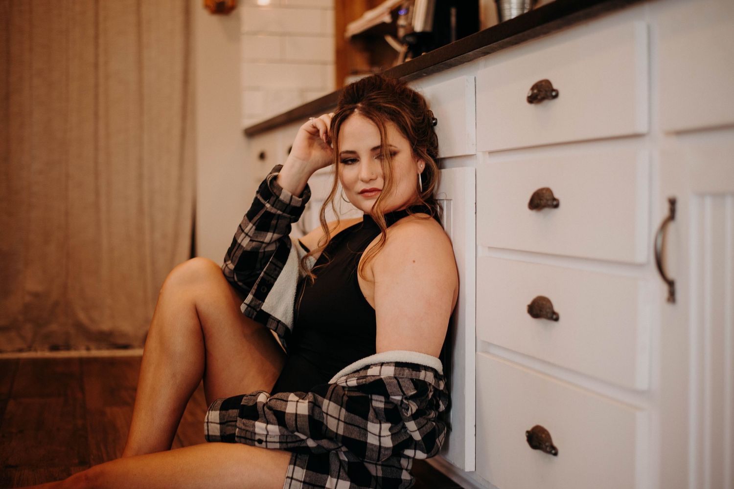 Young woman in plaid shirt and black top sitting on kitchen floor leaning against white dresser drawers.