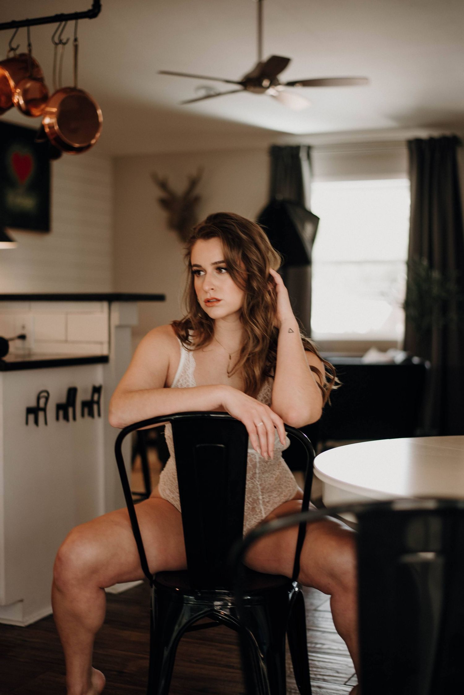 Young woman in lingerie sits on black metal chair in cozy kitchen with copper pots and ceiling fan.