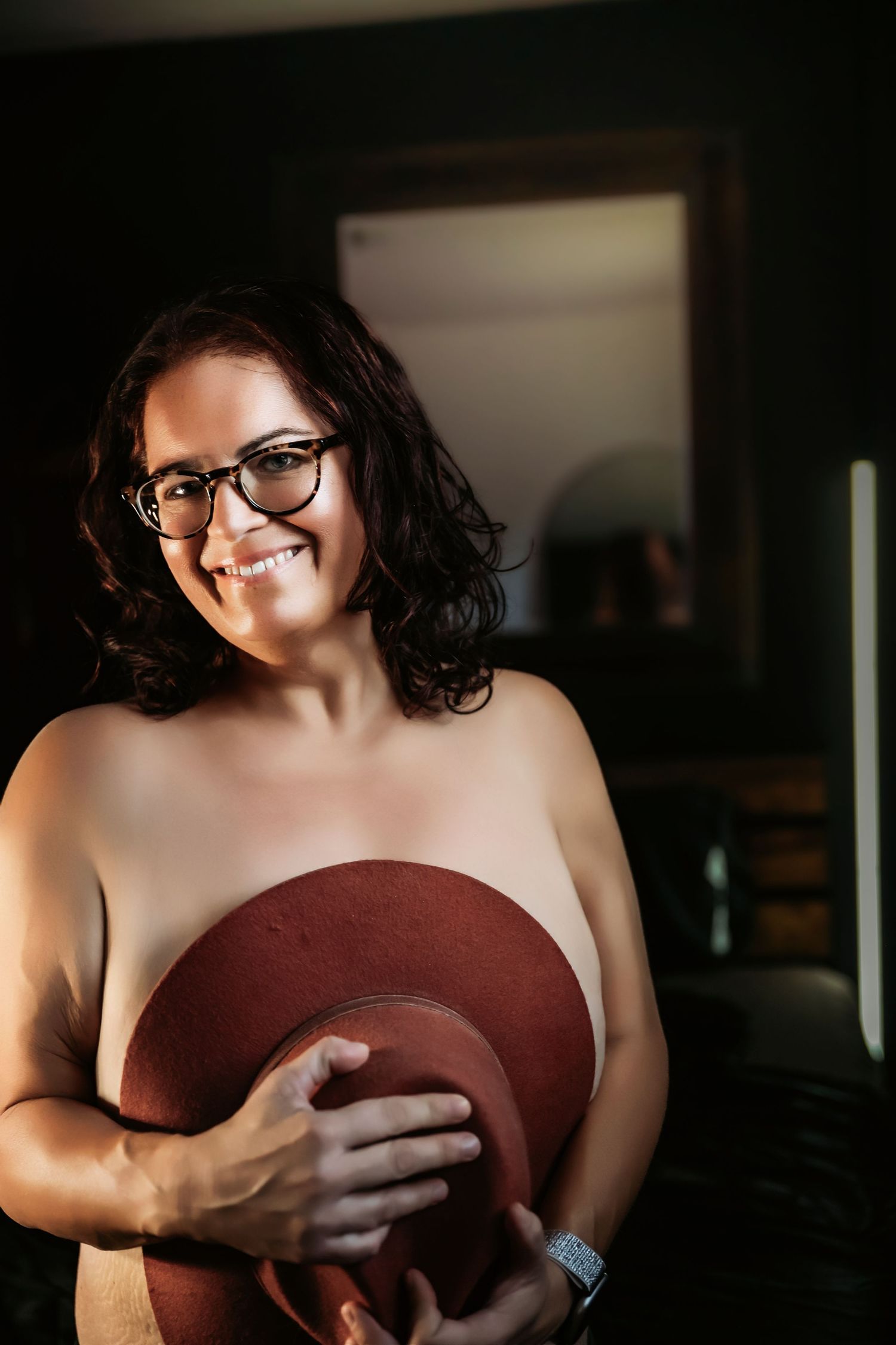 Smiling woman with glasses holding a brown hat in a warmly lit room.