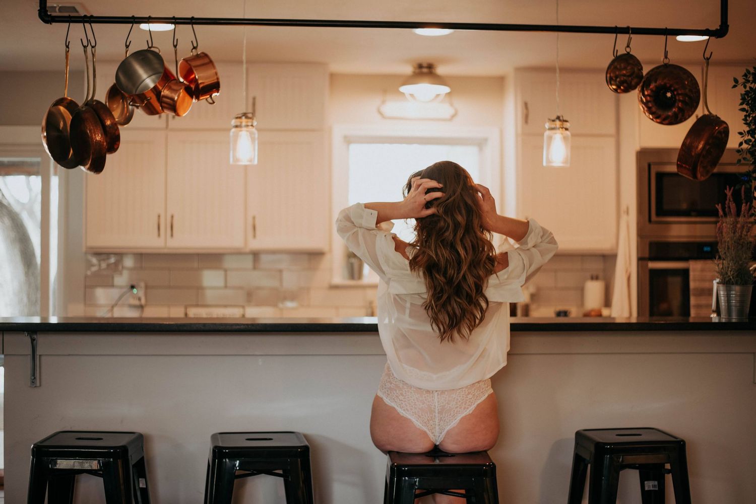 Woman in white shirt and underwear standing at kitchen counter with back to camera, hands in hair.