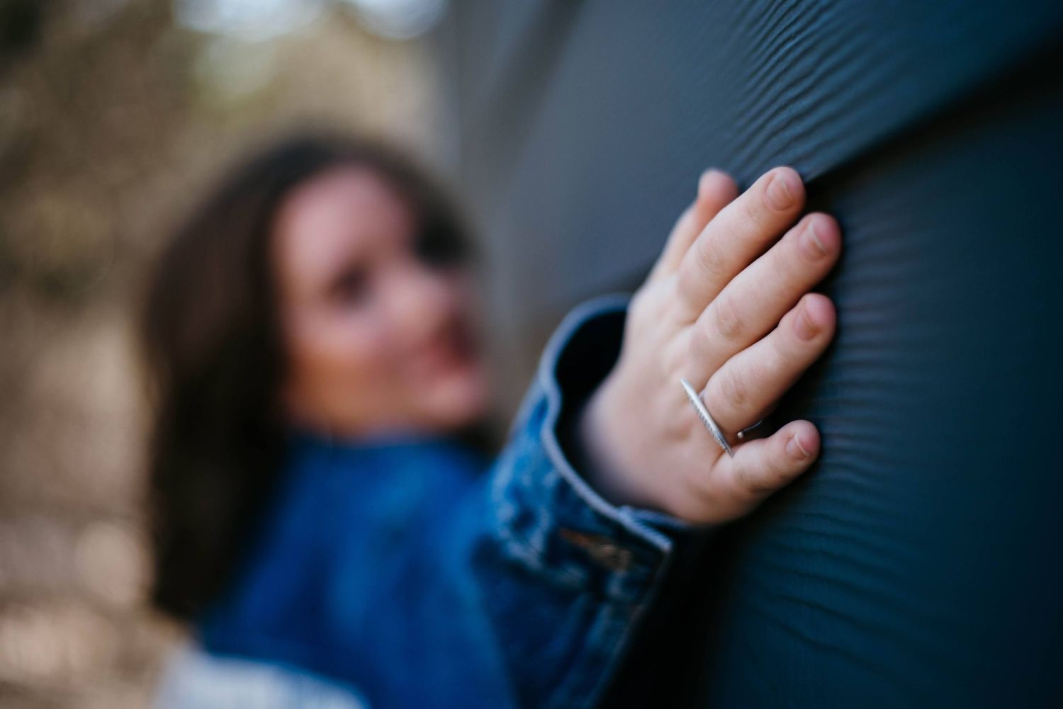 Close-up of woman's hand with silver ring touching gray corrugated wall wearing denim jacket.