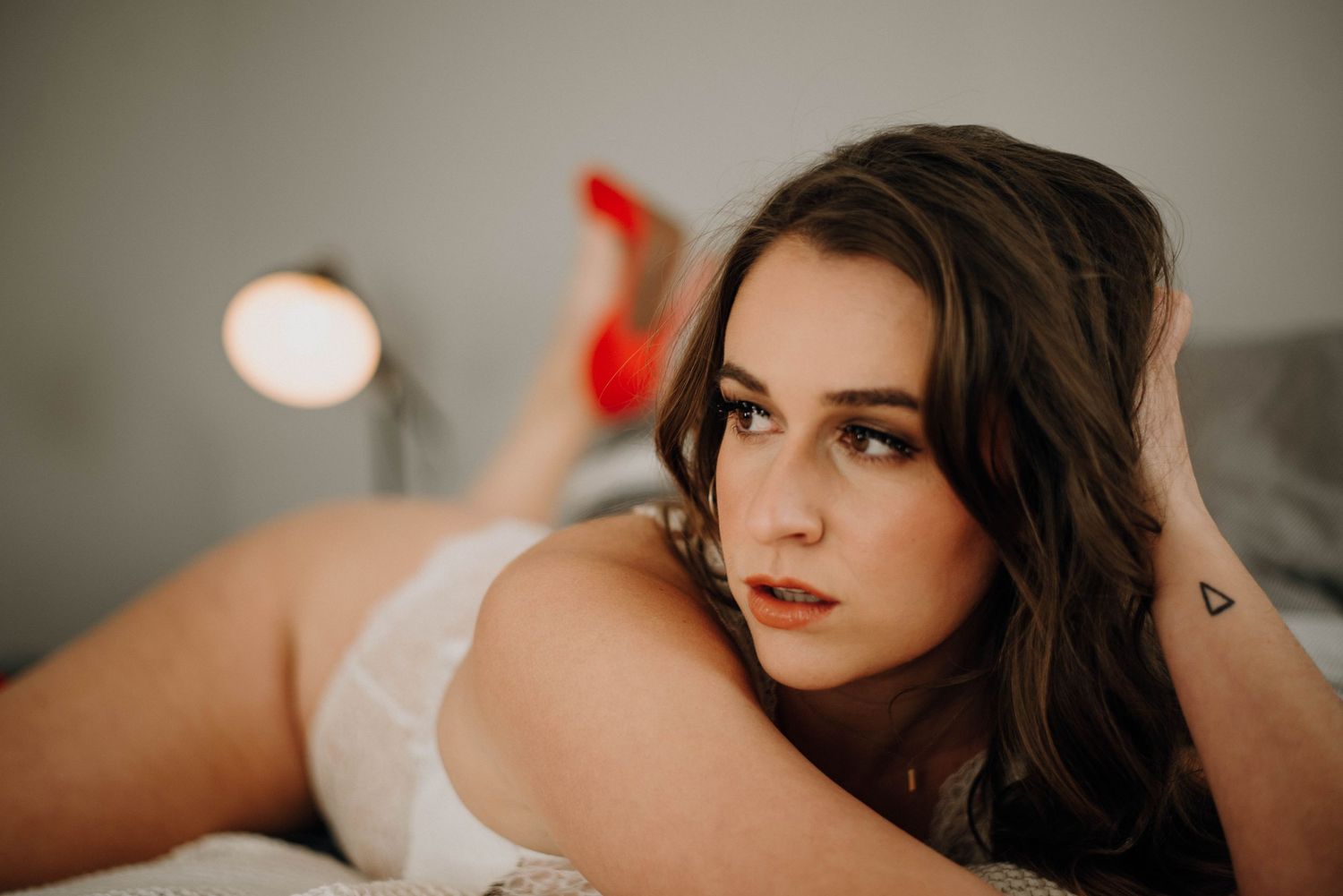 Young woman in white lingerie with red heels poses on a bed for a boudoir photo session.