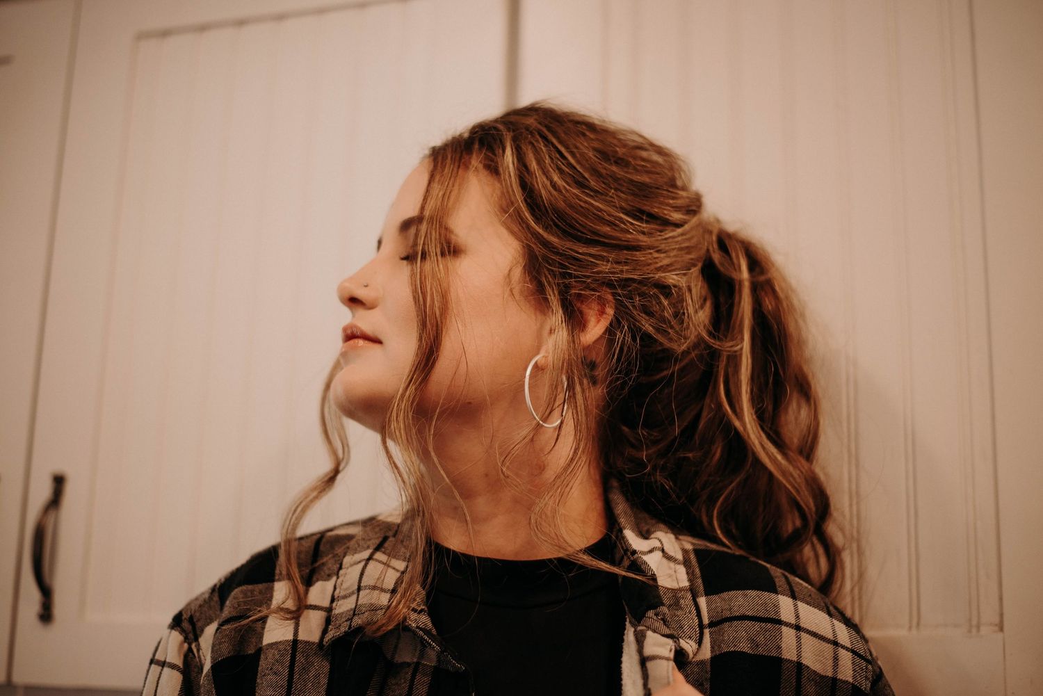 Young woman with curly updo hair and hoop earrings wearing plaid shirt, leaning against white cabinet.