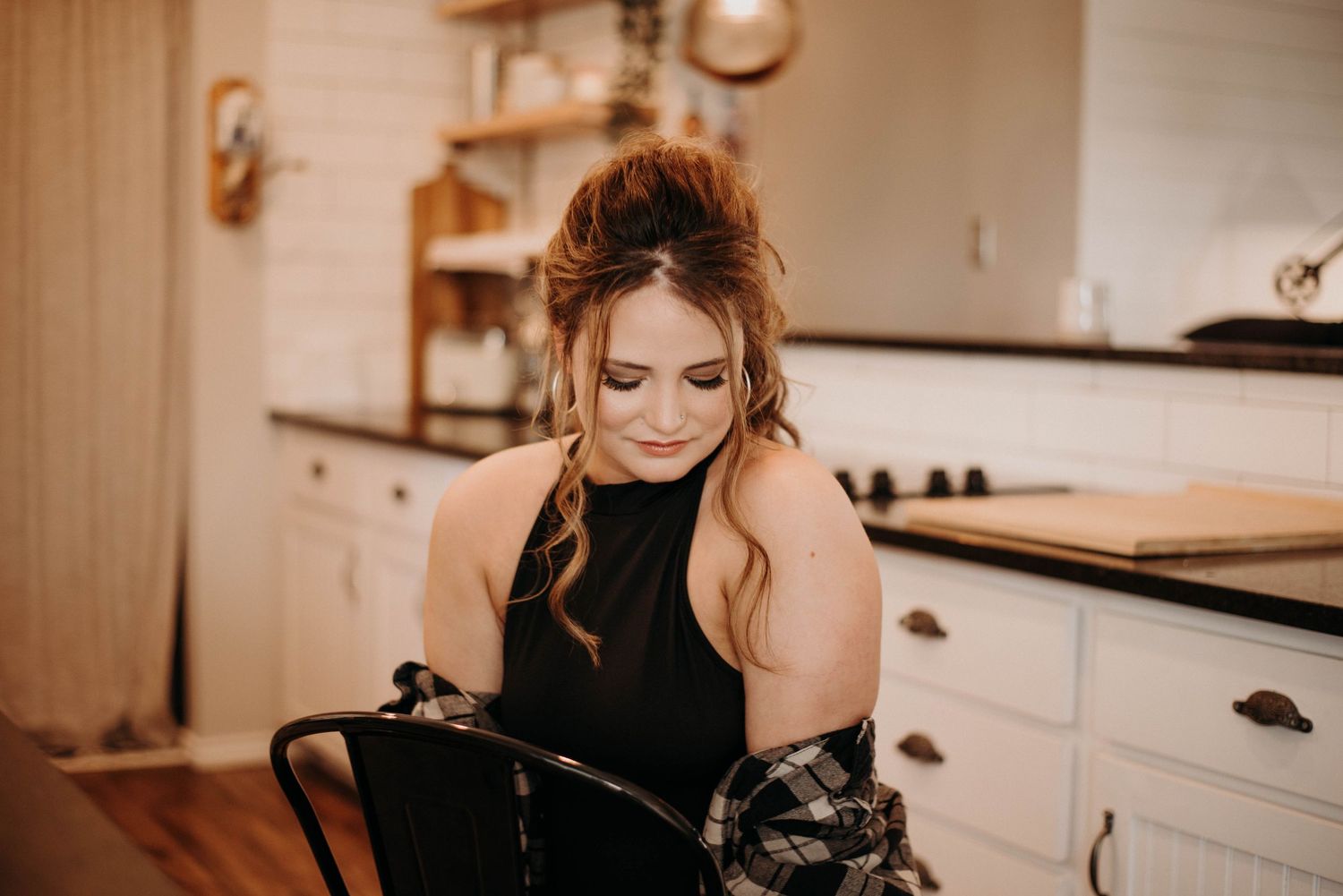 Young woman with updo hairstyle in black top and plaid shirt smiling in a cozy white kitchen.