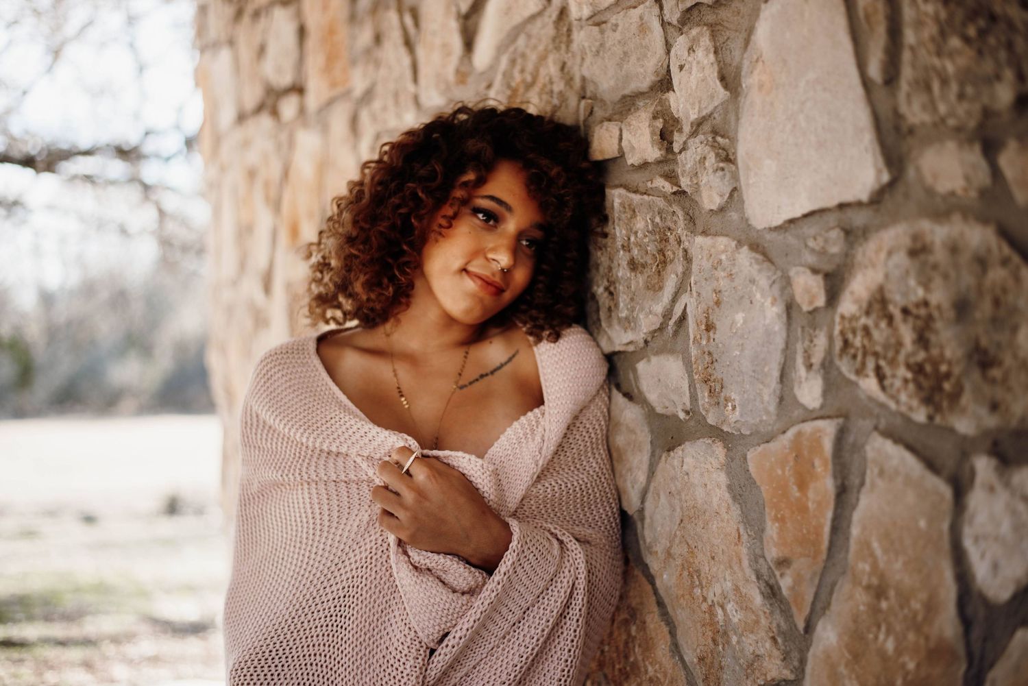 Young woman with curly hair wrapped in pink knit sweater leaning against rustic stone wall outdoors.