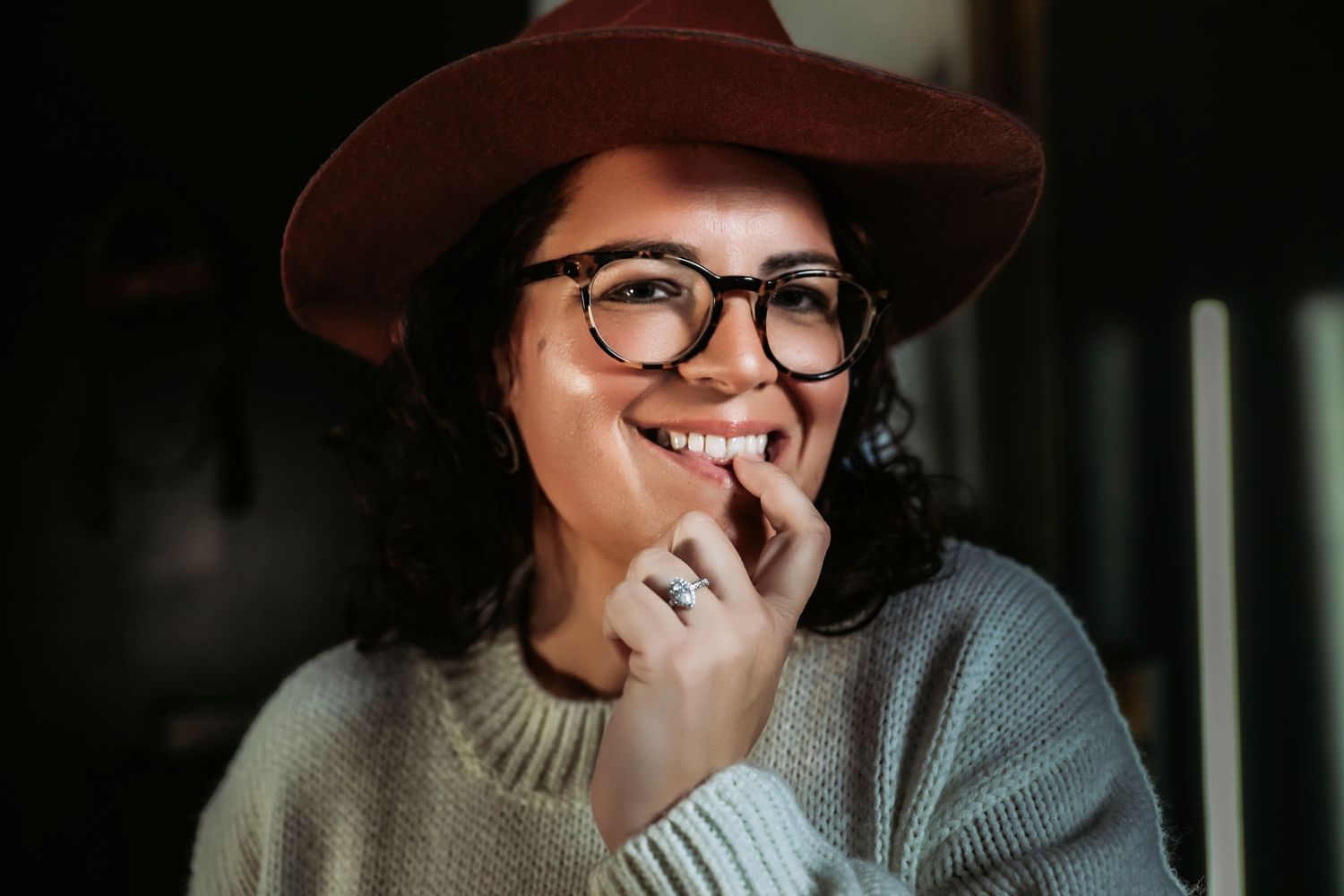 Smiling young woman wearing brown hat, black glasses, and knit sweater, posing cheerfully indoors.