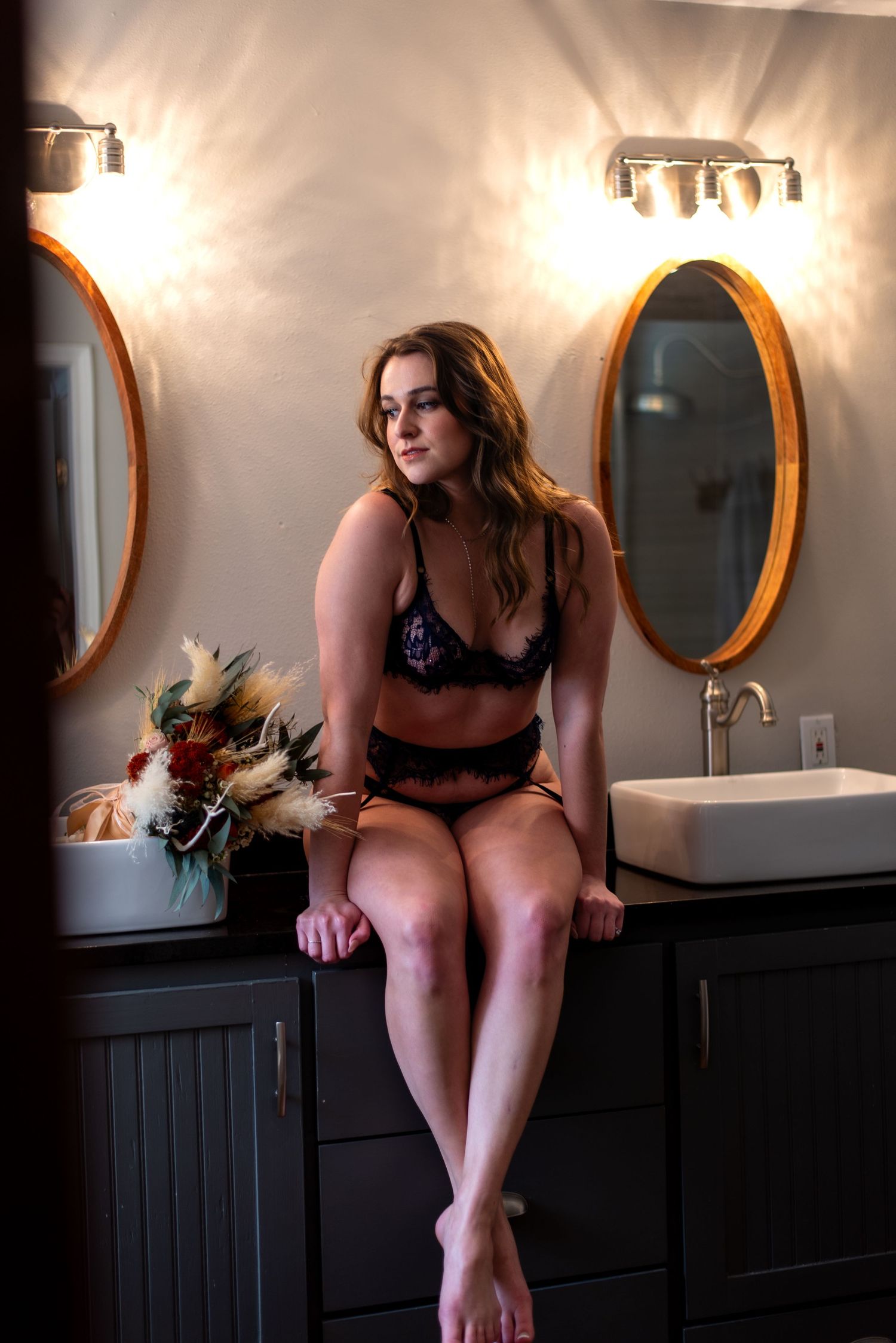 Woman in black lace lingerie sits on bathroom vanity counter beside oval mirrors and floral arrangement.