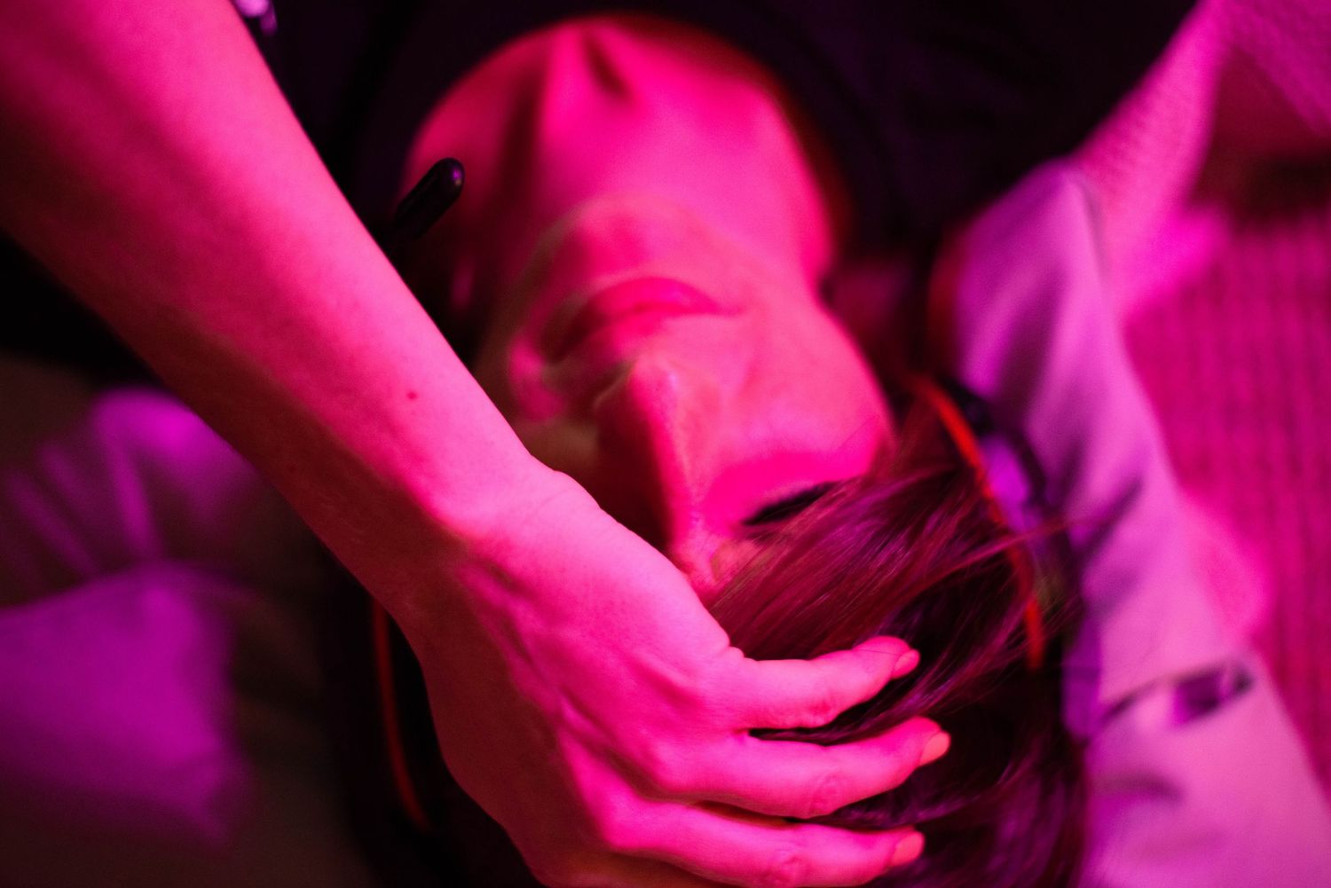 Close-up of hands gently cradling a womans head under pink and purple dramatic lighting.